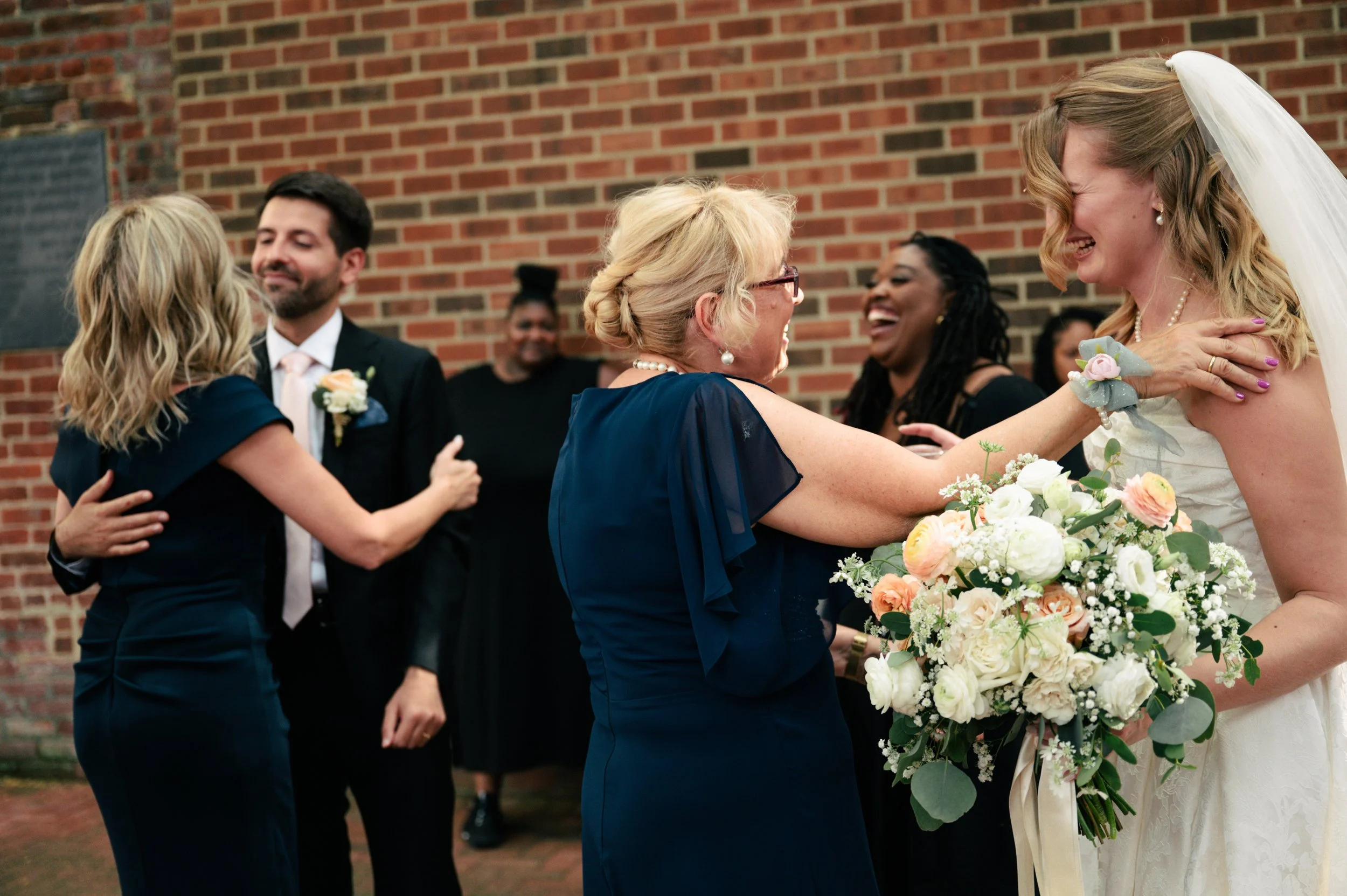 A bride and an older woman, likely her mother, share a joyful embrace at a wedding, with other guests smiling and dancing in the background against a brick wall.