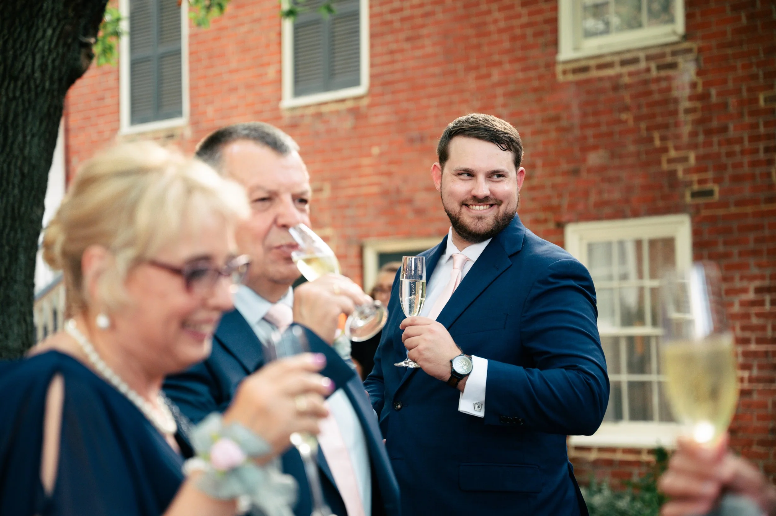 People dressed in formal attire celebrating outdoors with drinks in hand, smiling, with a brick building in the background.
