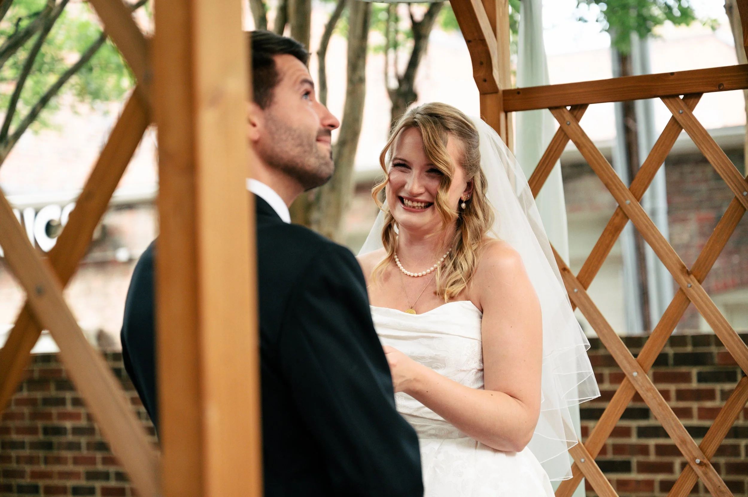 A bride and groom standing close together, smiling during their wedding ceremony outdoors, with a wooden lattice structure and trees in the background.