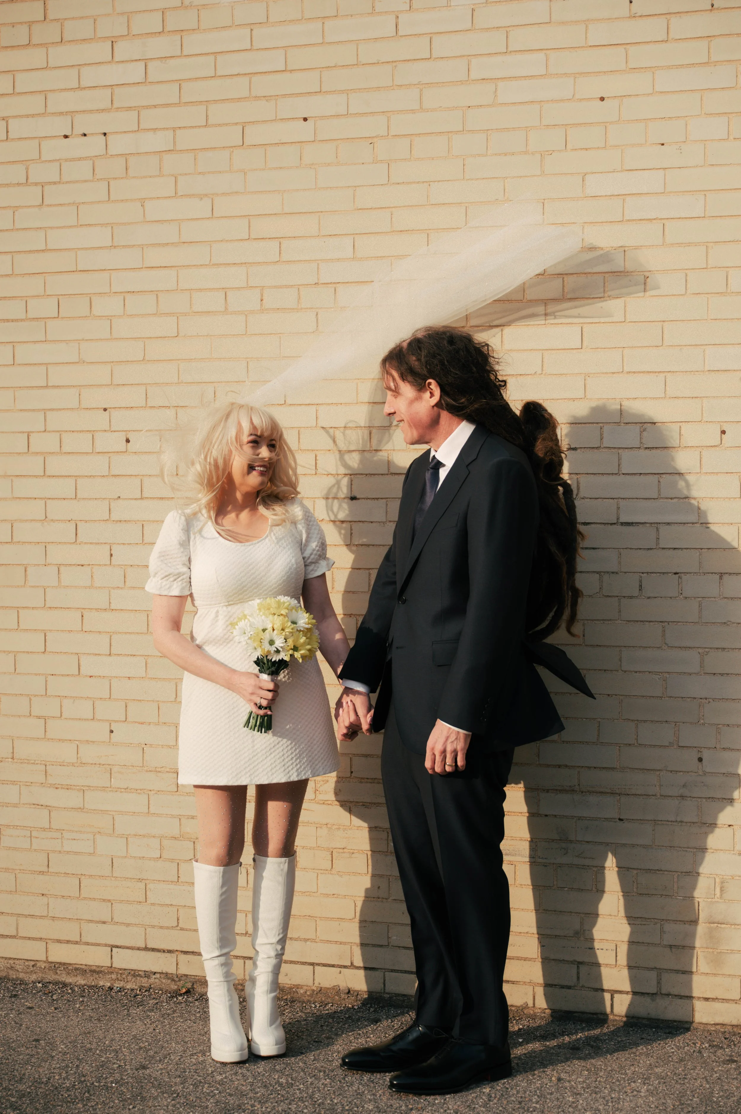 A woman in a white dress and knee-high white boots holding a bouquet of white flowers, smiling at a man in a black suit who is holding her hand and smiling back. They are standing against a beige brick wall, appearing happy on a sunny day.