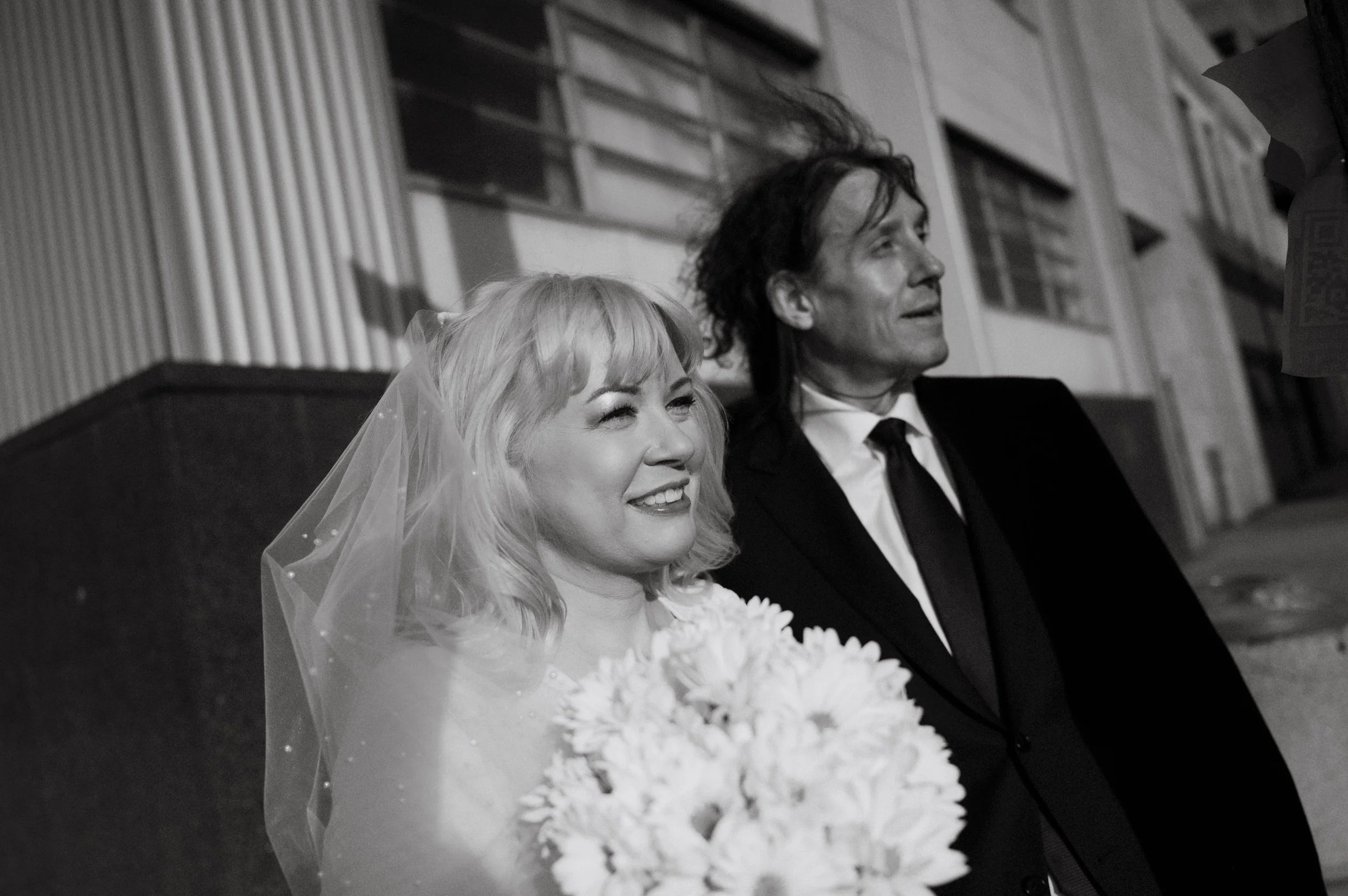 A bride and groom sitting together at their wedding, smiling. The bride has blonde hair with a veil and is holding a bouquet of flowers. The groom has dark, curly hair and is dressed in a suit with a tie. They are seated indoors with a wood-paneled w