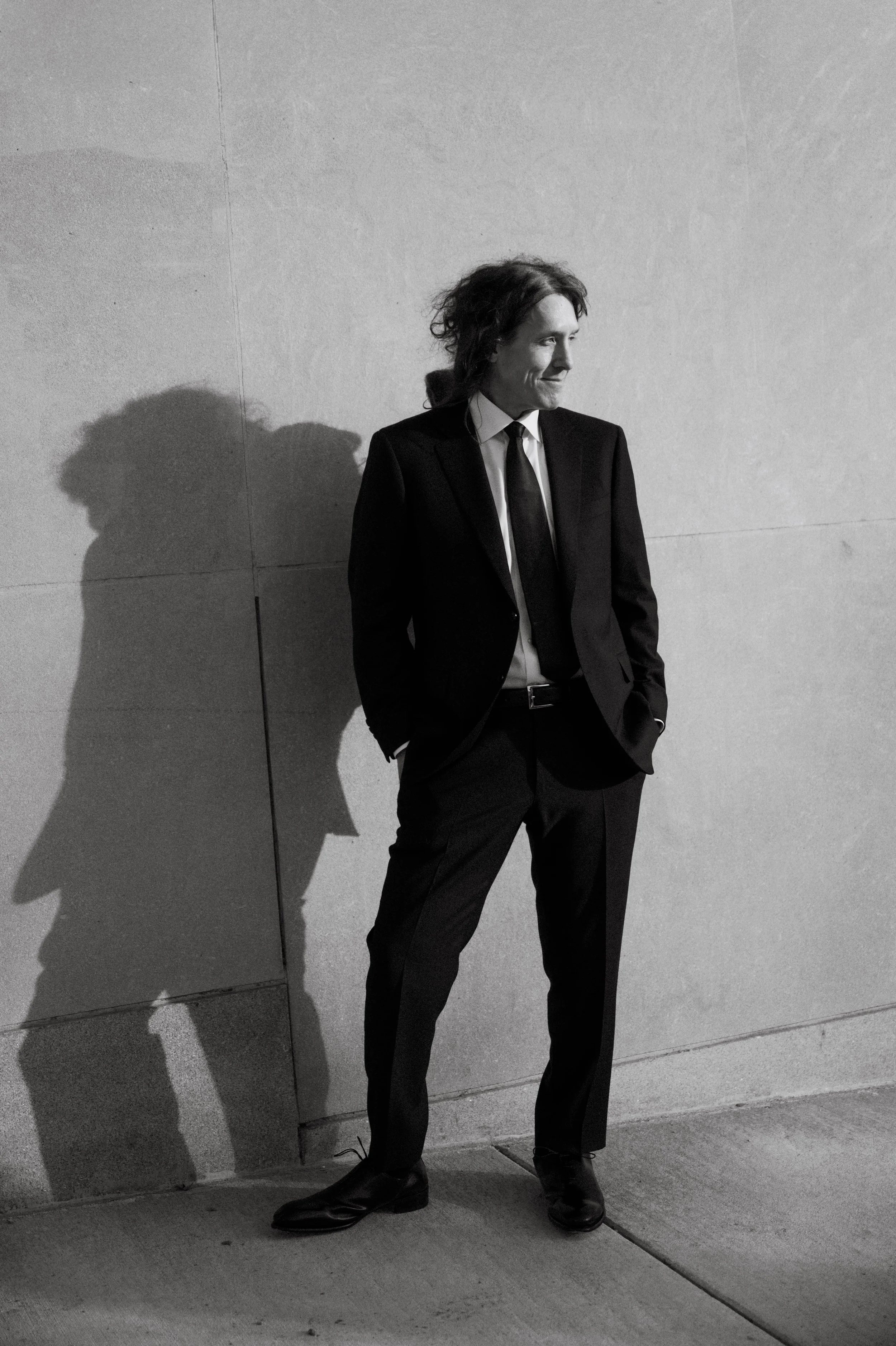 Black and white photo of a man with curly hair wearing a suit, standing against a plain wall, with his hands in his pockets.