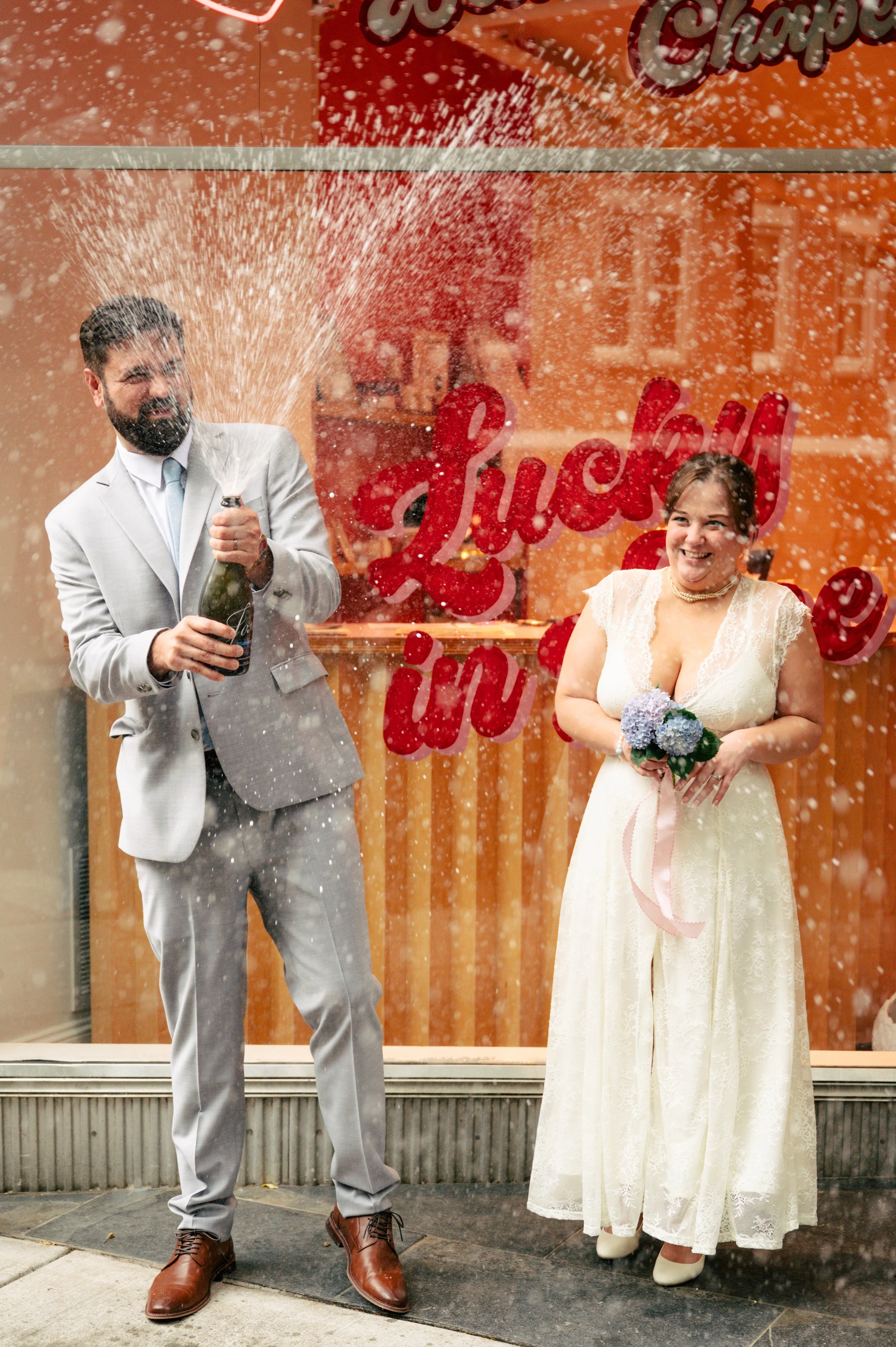 A man in a light gray suit and brown shoes is opening a bottle of champagne, causing it to spray out foam. Next to him, a woman in a white lace dress and white shoes holds a bouquet of flowers and smiles. They are standing in front of a window and other decorative writing and balloons in the background.