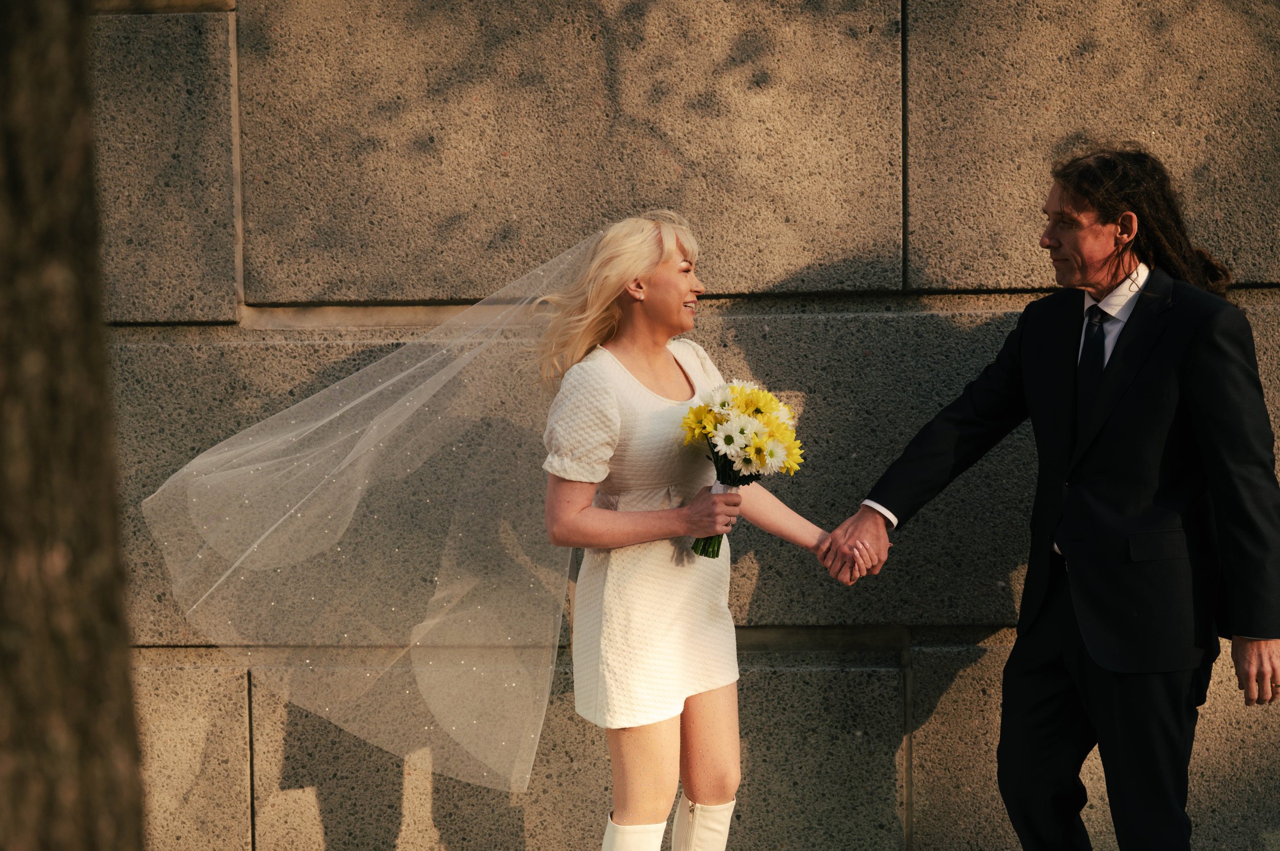 A bride wearing a white dress and veil holding a bouquet of yellow and white flowers, smiling at a man in a black suit while holding hands against a stone wall, during a wedding ceremony.