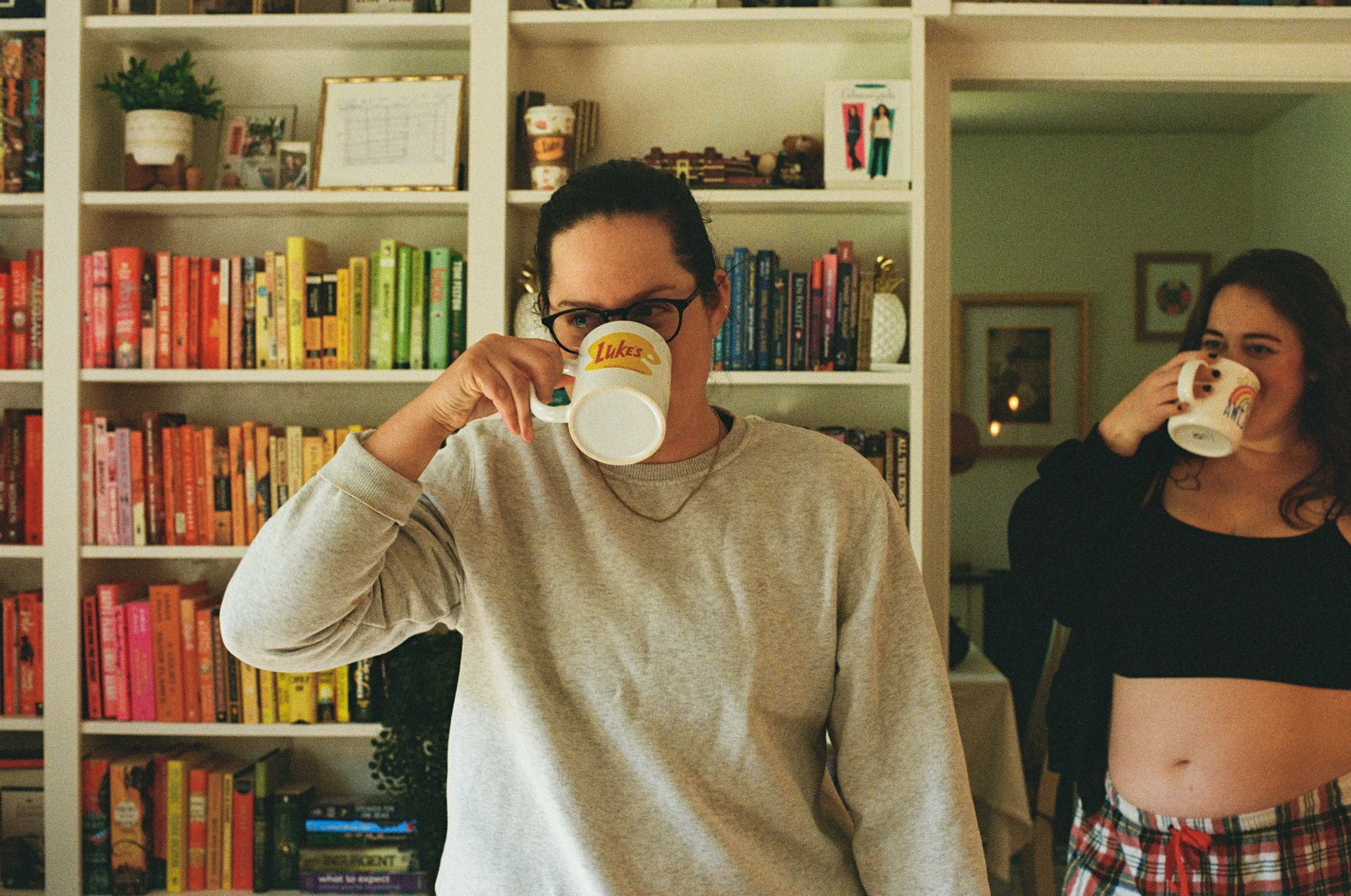 Two women drinking from mugs in a room with a bookshelf and framed pictures. The woman on the left has glasses and curly hair, wearing a beige sweater. The woman on the right has dark hair, wearing a black top and plaid shorts.