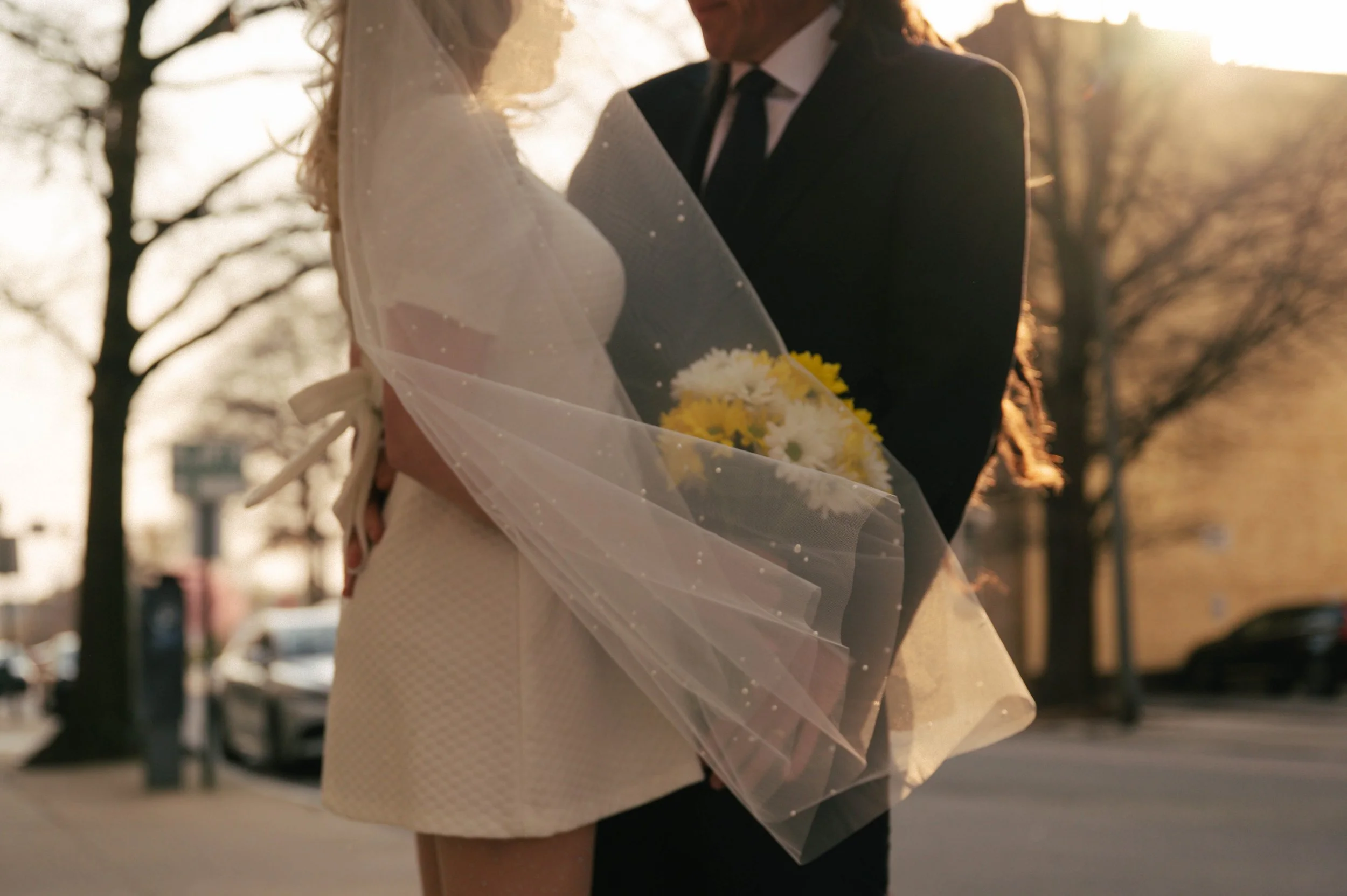 A couple dressed in formal wedding attire, standing close together outdoors during sunset, with the bride holding a bouquet of white and yellow flowers wrapped in sheer fabric.