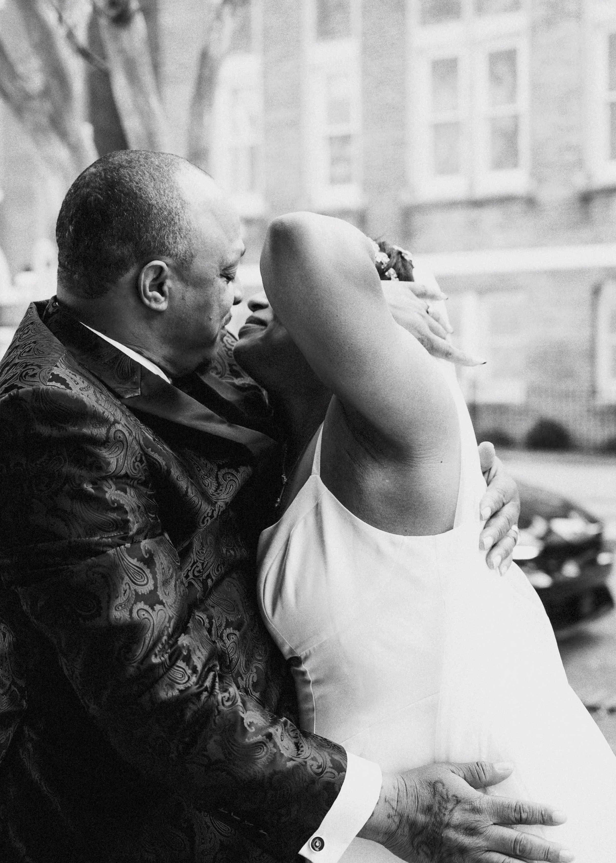 An older man and a woman are sharing an intimate moment, with their faces close and their foreheads touching, in front of a window with a cityscape background.