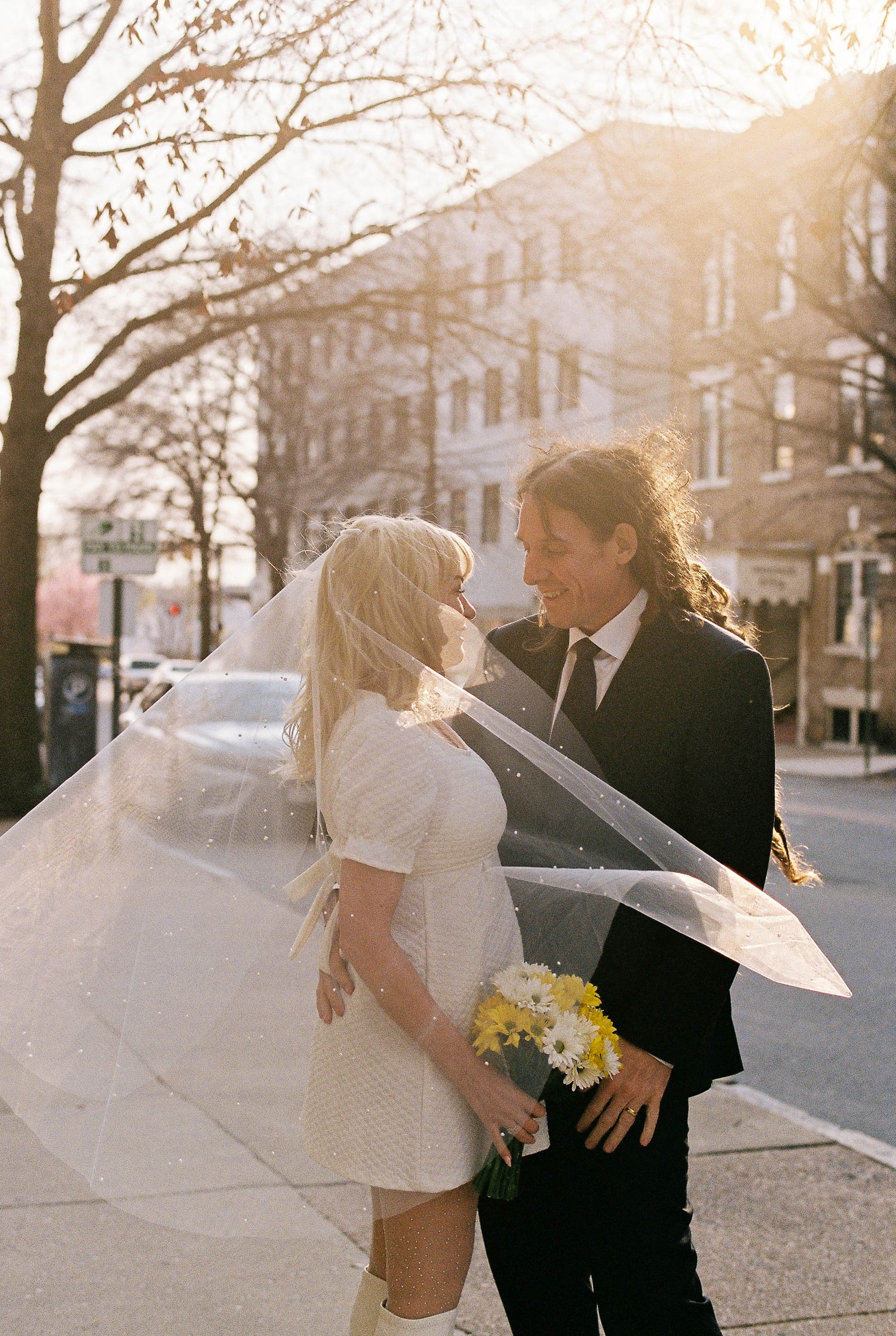 A woman in a wedding dress and a man in a suit are standing close together on a city street, smiling at each other with the sun shining behind them. The woman is holding a bouquet of yellow and white flowers, and a veil is draped over her shoulders.
