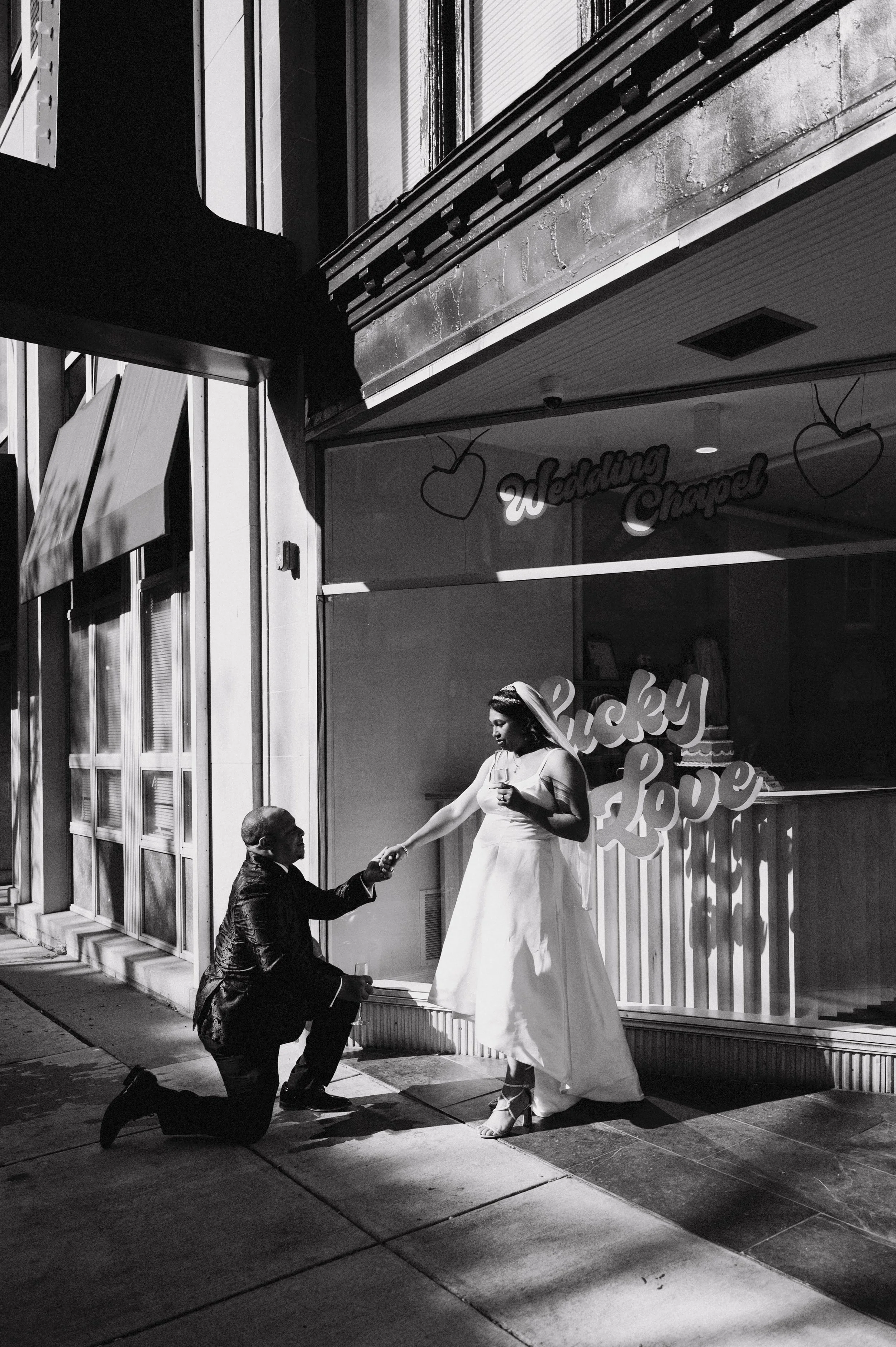 A woman in a white dress and veil is standing outside a storefront, handing a glass of champagne to a man kneeling in front of her. The storefront has signs that say 'Wedding Chapel' and features hearts and decorative lettering.