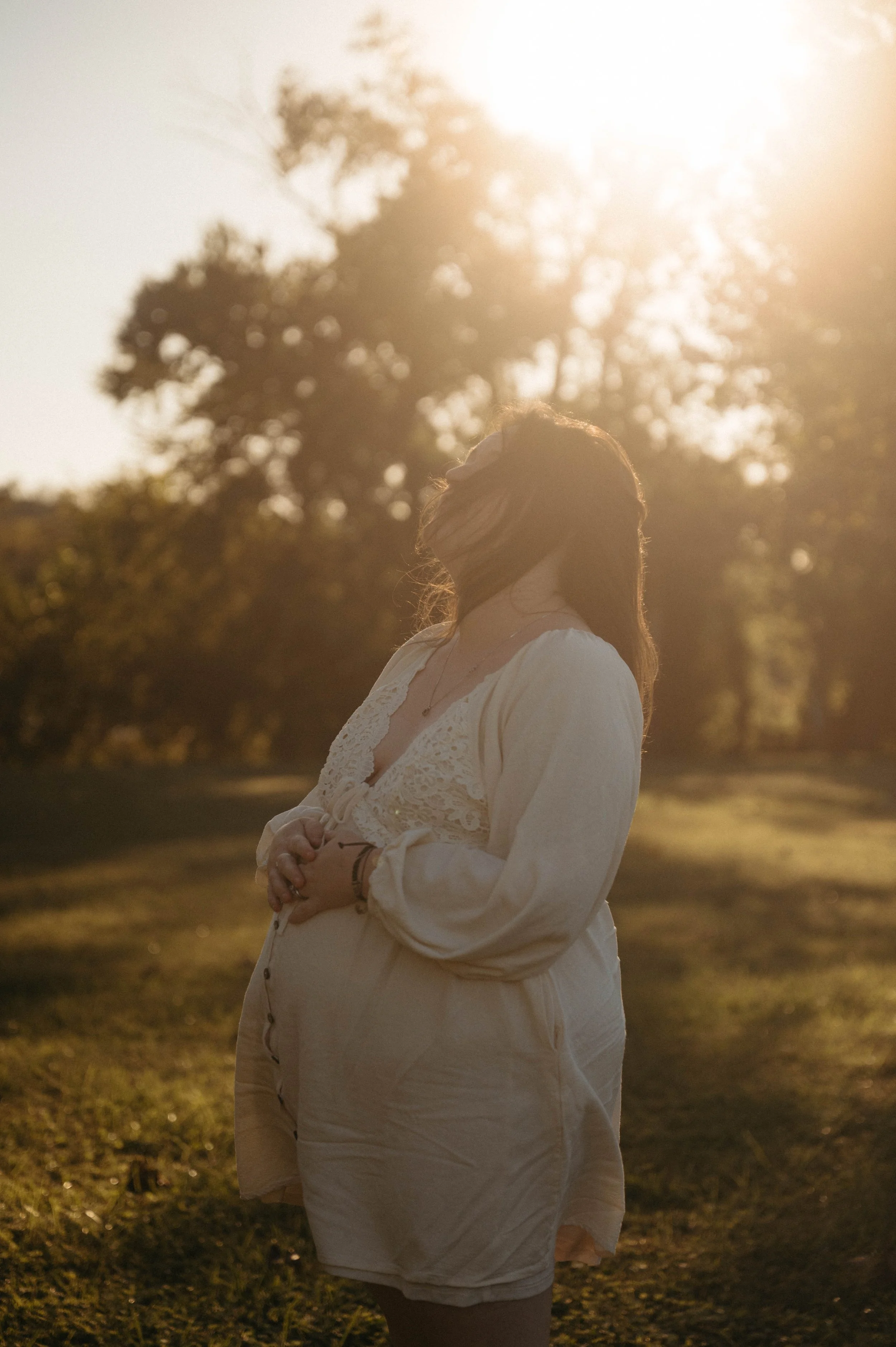 A woman standing outdoors in the late afternoon sun, with her eyes closed and head tilted back, wearing a white, lacy dress. She has long, wavy hair and is surrounded by trees and grass and is embracing her pregnant belly.