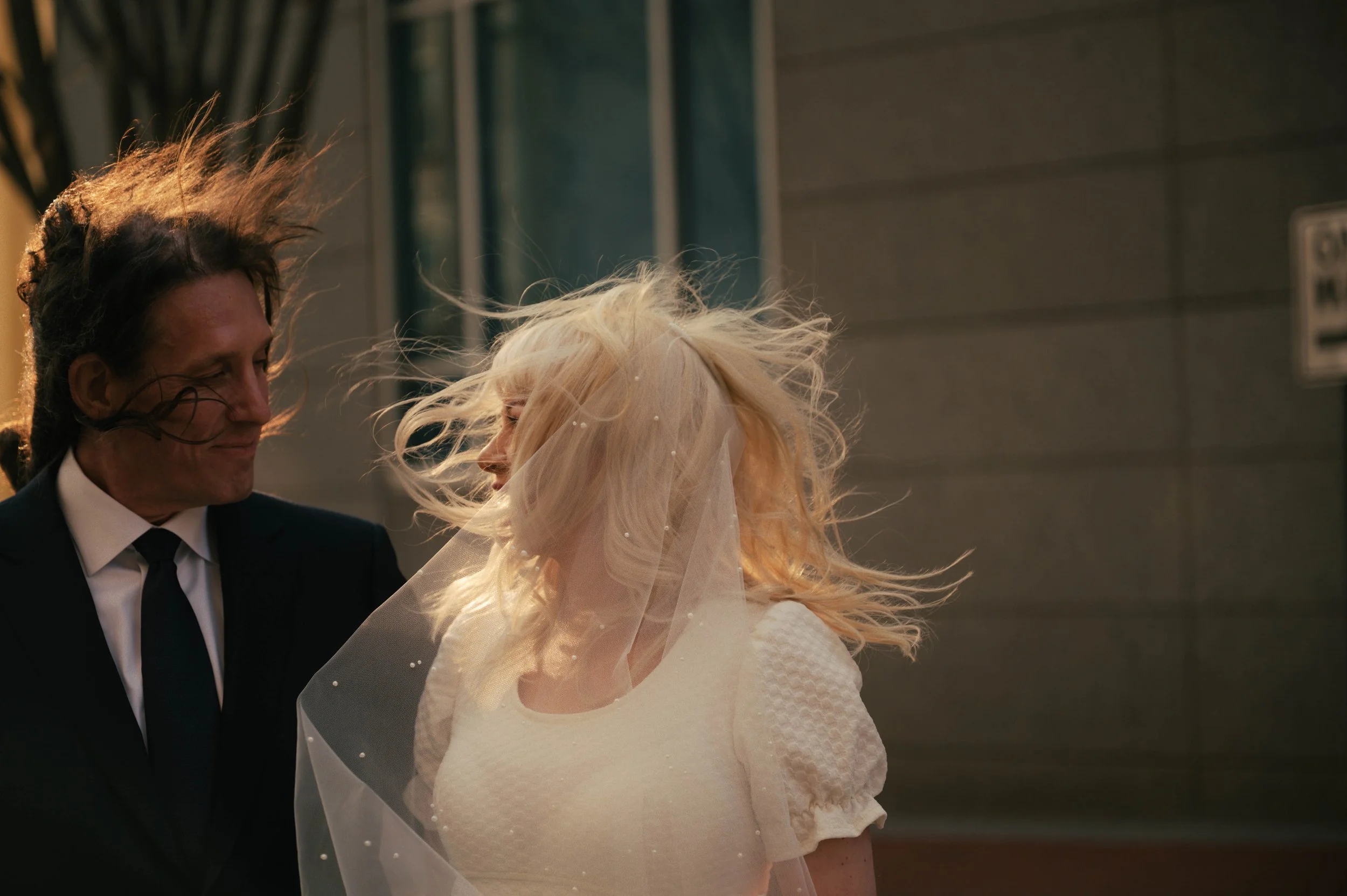 A man and a woman, possibly at a wedding, are dancing outdoors at night with wind blowing their hair. The man is wearing a black suit and tie, and the woman is wearing a white dress and veil.