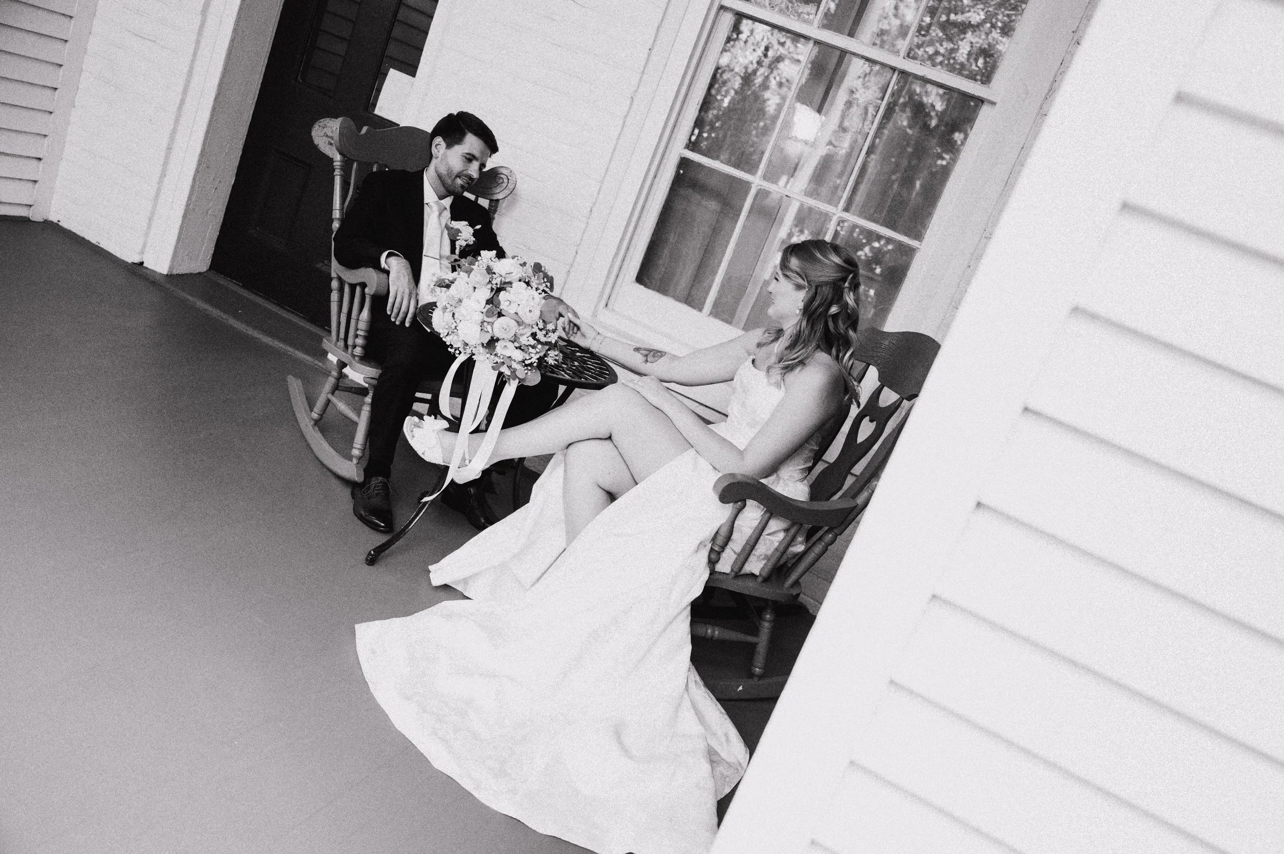 Black and white photo of a man and woman sitting on rocking chairs, holding hands, with the man smiling and looking at the woman, on a porch with large windows, decorated with flowers.