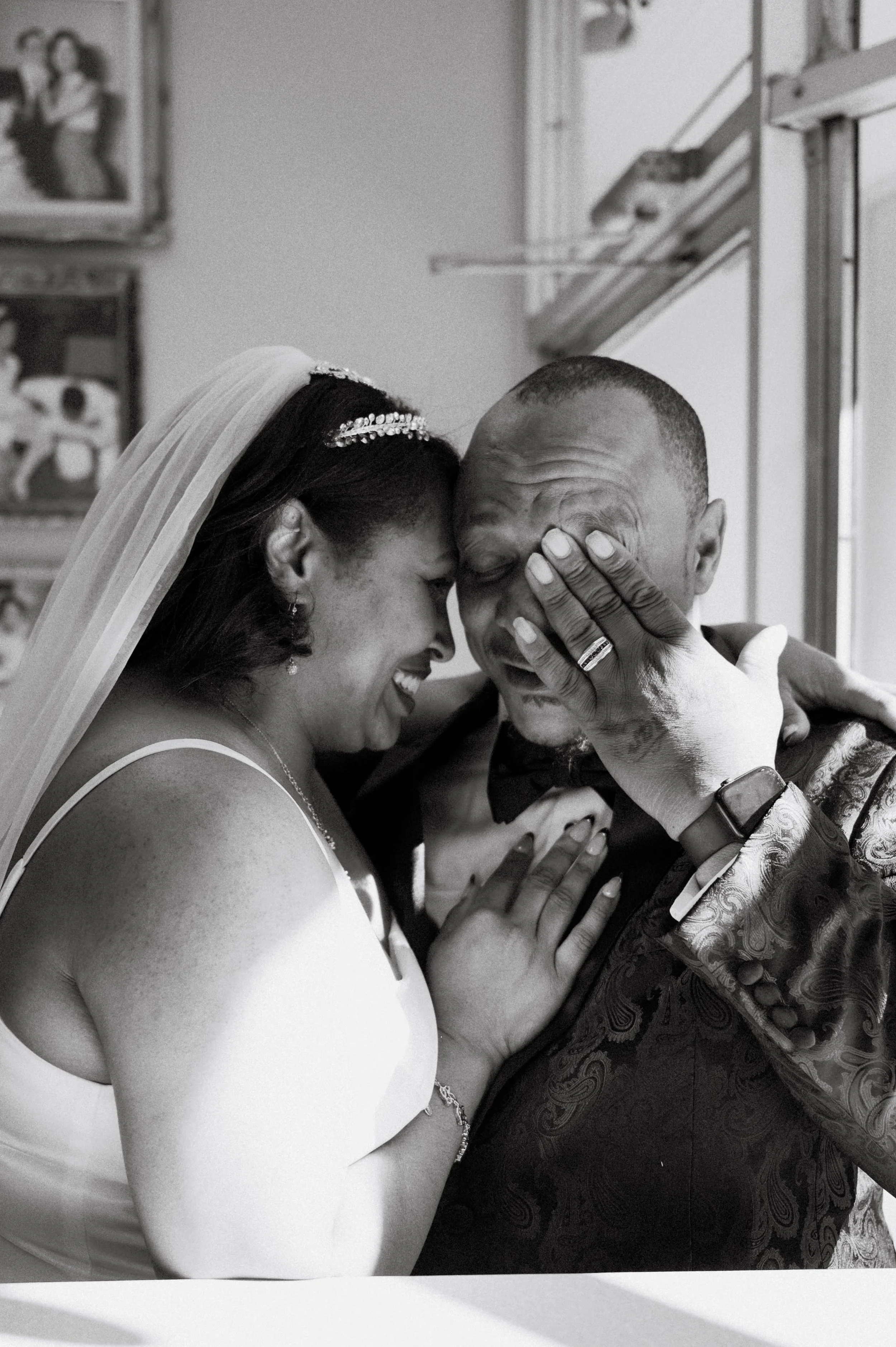 In this black and white photo, a bride and groom are sharing an intimate moment, touching foreheads and smiling. The bride is wearing a veil and a sleeveless dress, and the groom is in an ornate suit jacket. Both are visibly happy and close, with their hands touching. The background features some framed photographs and a window.