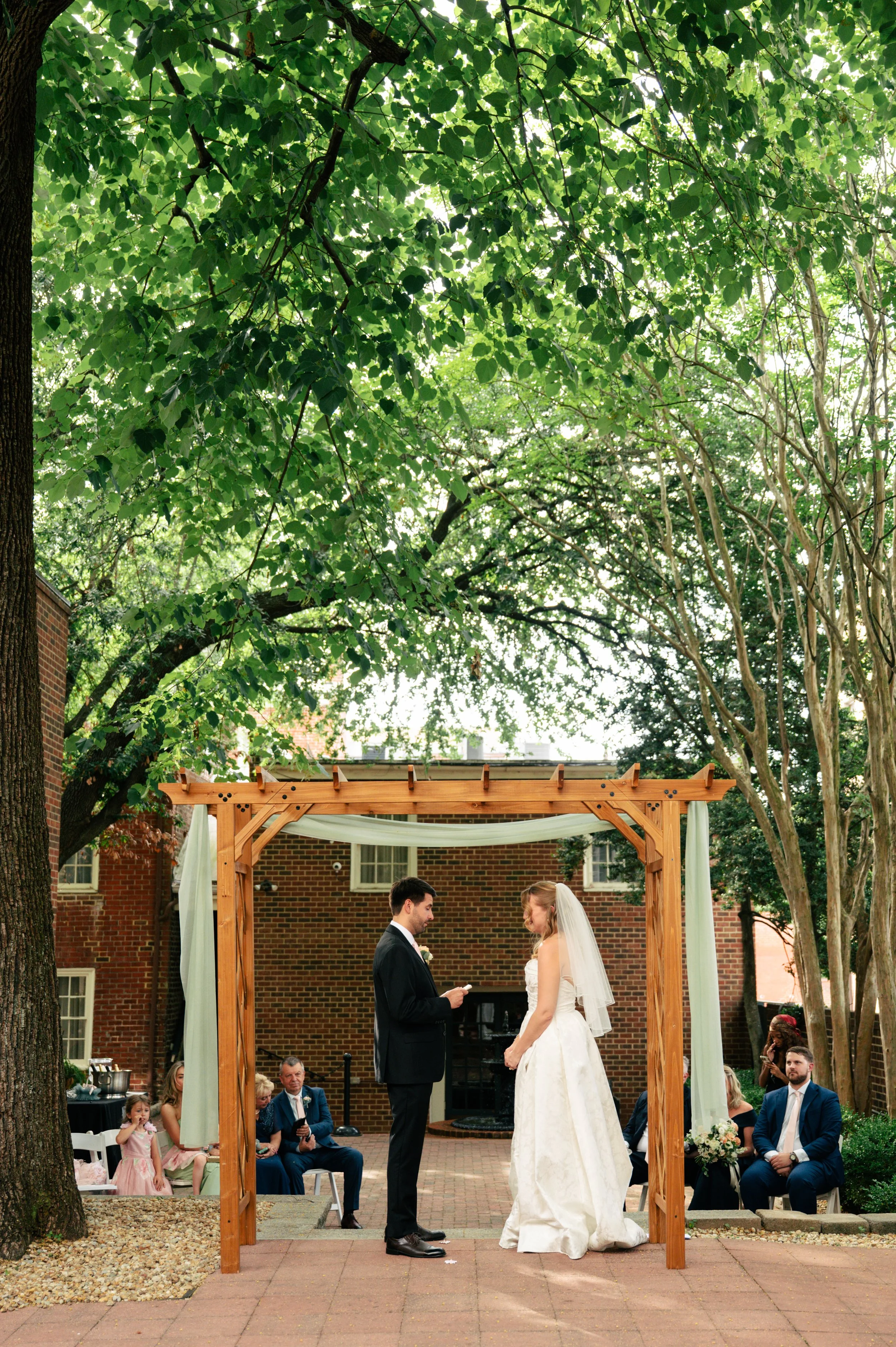 A wedding ceremony taking place outdoors under a wooden arbor with white drapes, with the bride and groom standing face to face, exchanging vows, surrounded by seated guests and children, with large leafy trees and a brick building in the background.