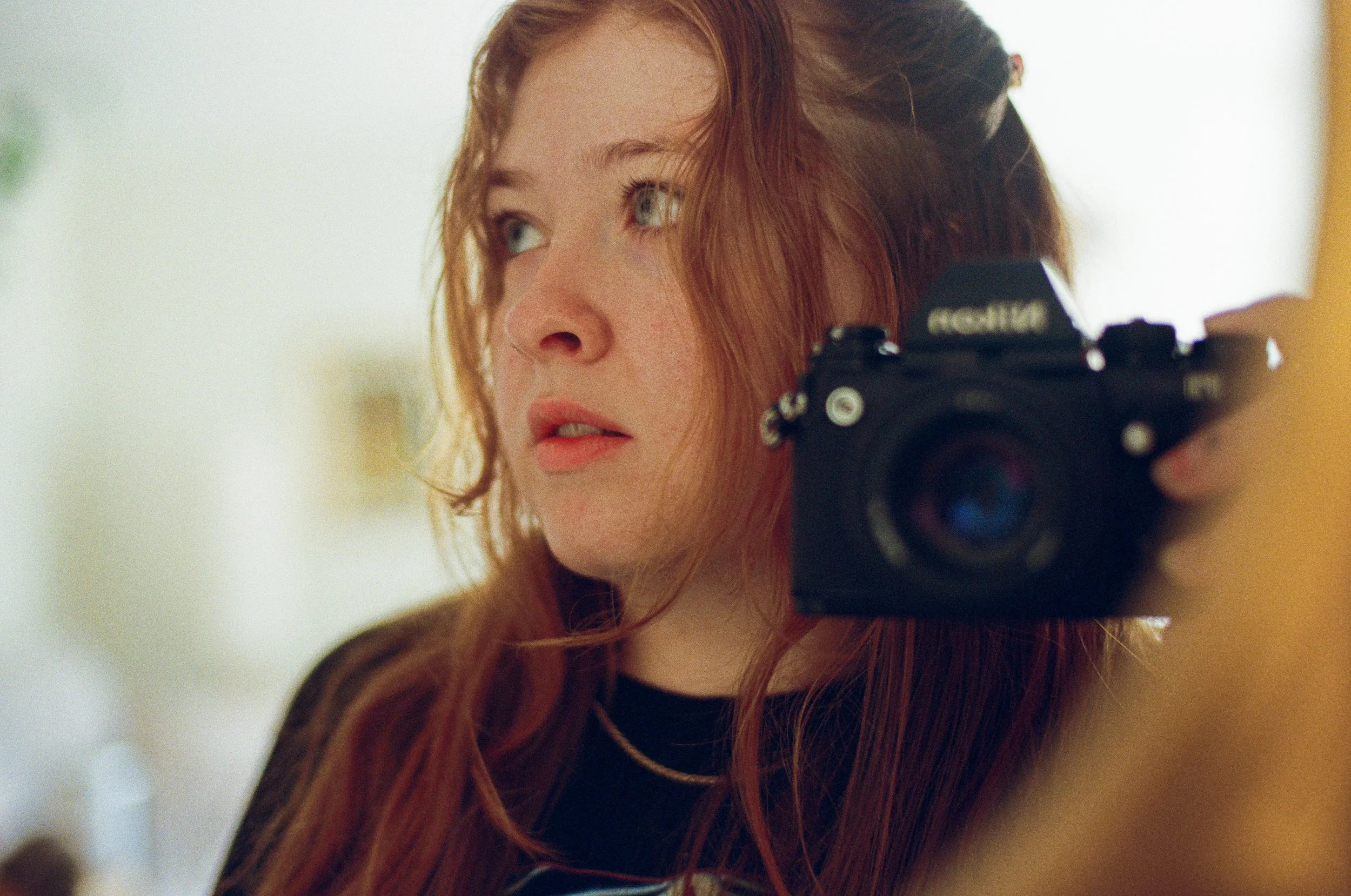 A young woman with red hair holding a camera to her shoulder, looking to the side.