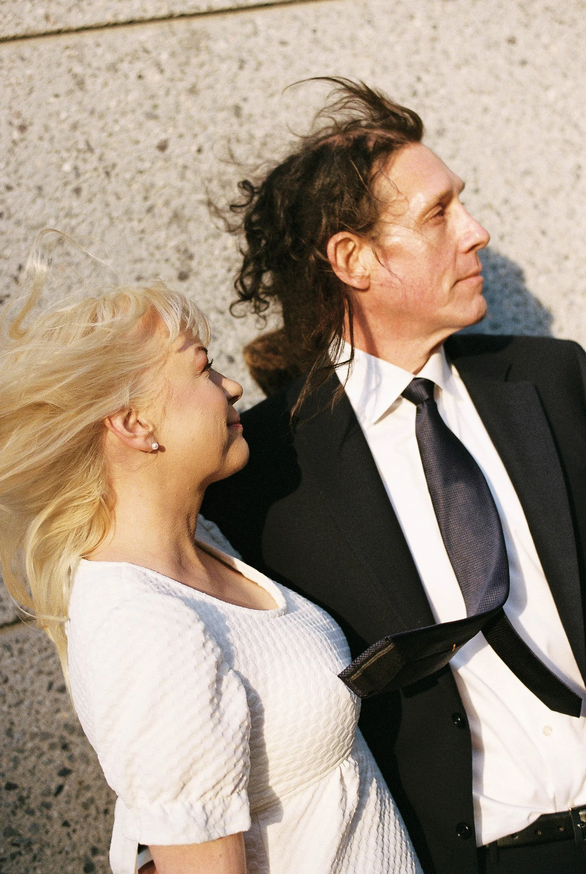 A man and a woman lying on a gravel surface, side by side, with the woman facing upward and the man facing sideways. The woman has long, blonde hair and a white textured dress. The man has dark, curly hair, a black suit, white shirt, and black tie.