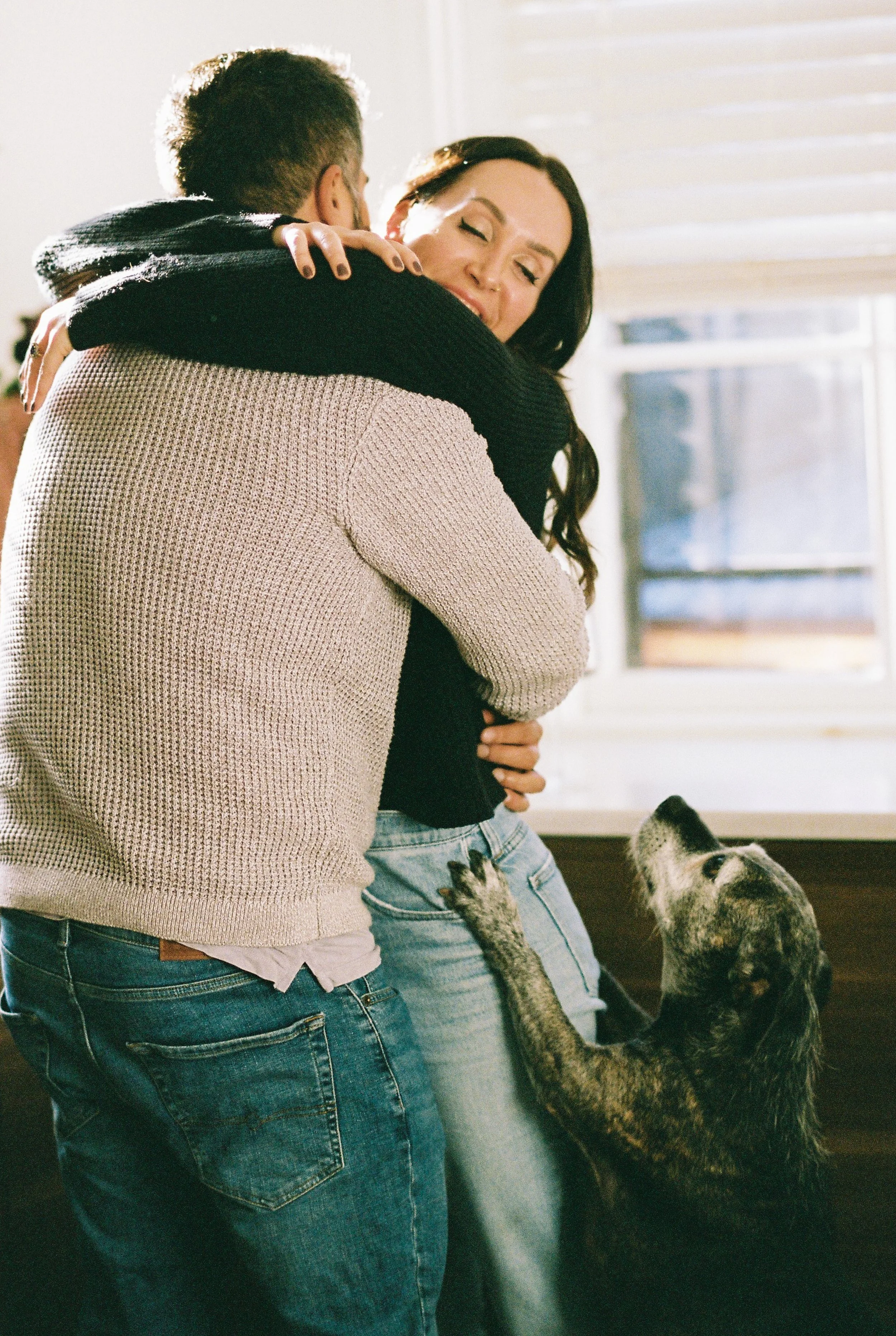 A woman and a man hug indoors, with a puppy reaching up to them.