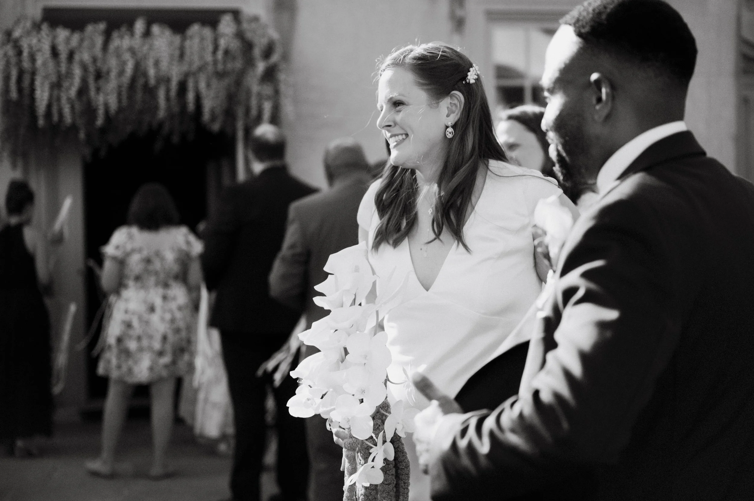 A smiling woman in a wedding dress and earrings, holding a bouquet of white flowers, standing next to a man in a tuxedo, during a wedding ceremony in a decorated indoor venue.