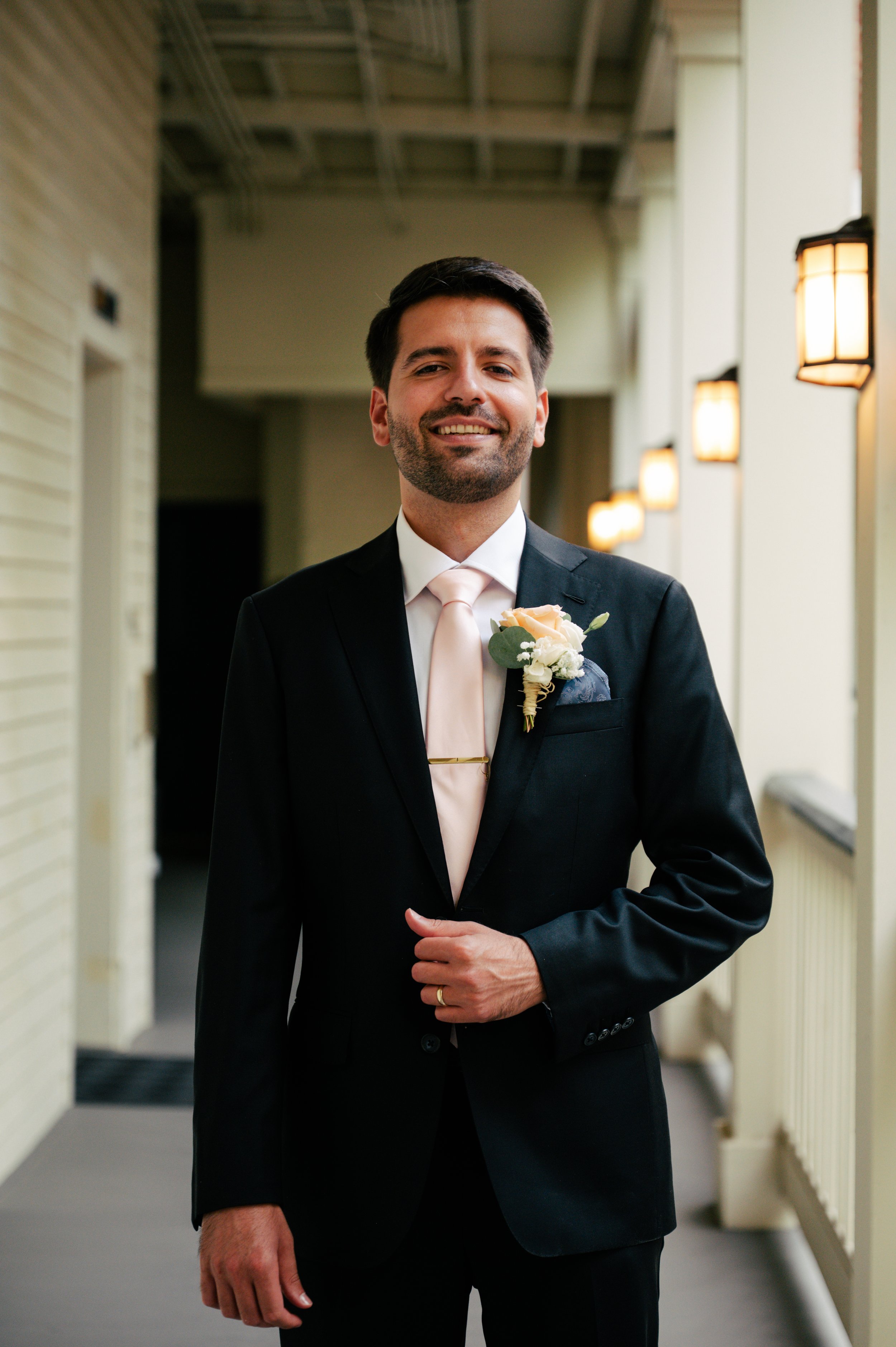 A man dressed in a black suit, white shirt, and light pink tie, smiling and standing in a hallway with wall-mounted lights.