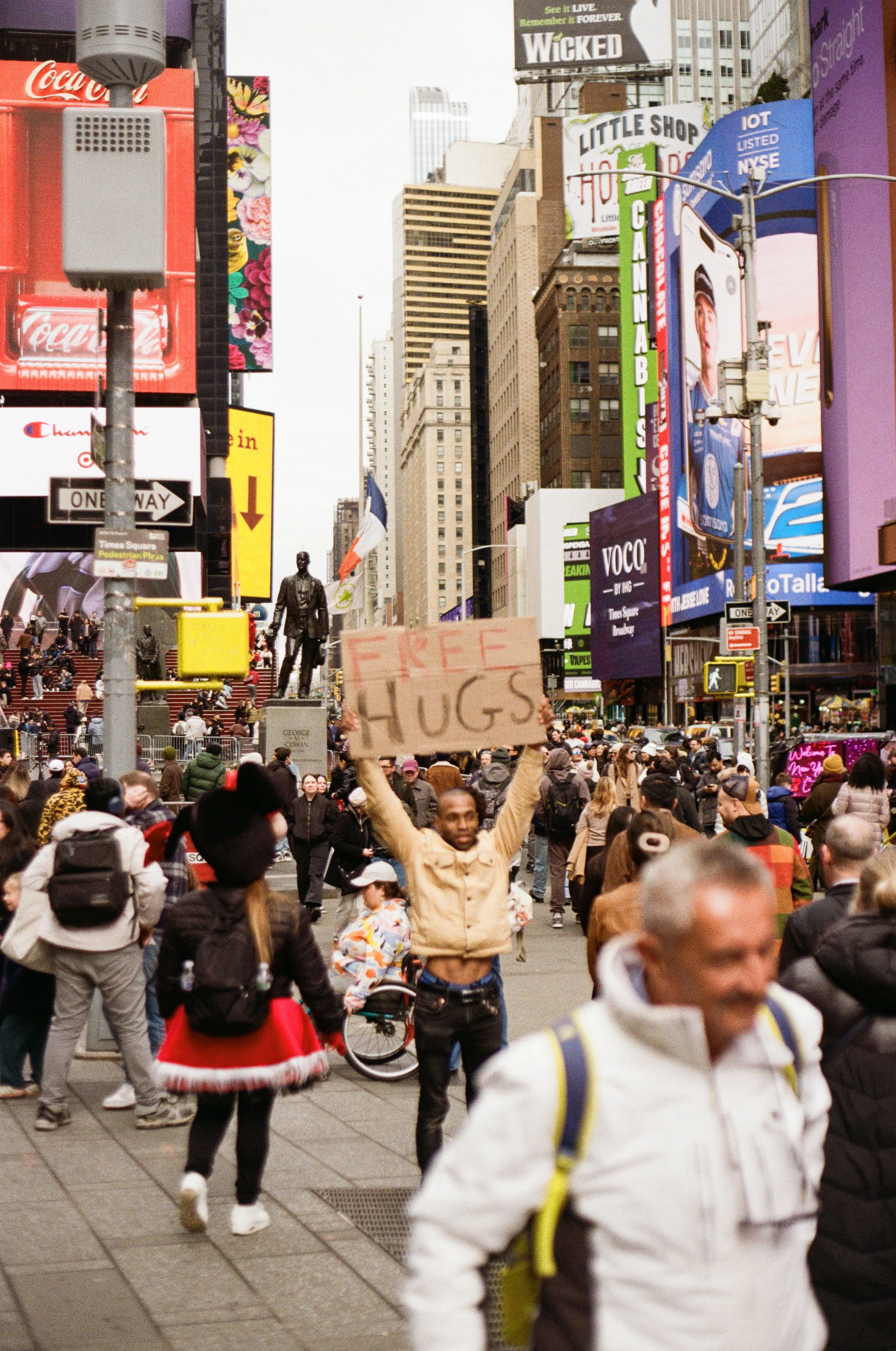 A crowded city street scene during daytime with people walking and a person holding a cardboard sign that says 'FREE HUGS' in the foreground. High-rise buildings, billboards, and signs are visible in the background, indicating a busy urban area.