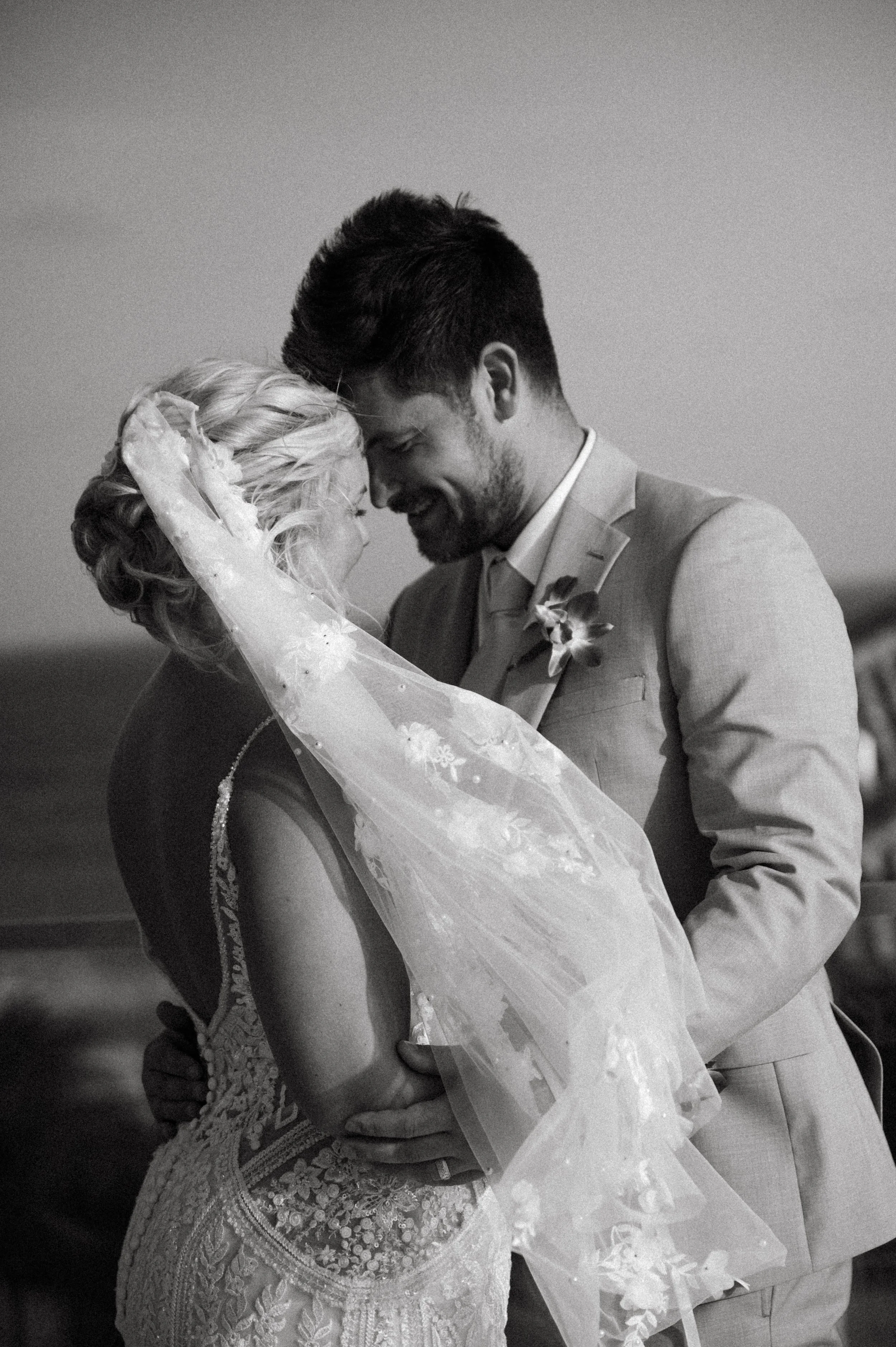 A black-and-white photo of a bride and groom on their wedding day, touching foreheads and smiling, with the groom wearing a light-colored suit and the bride in a lace wedding dress with a veil.