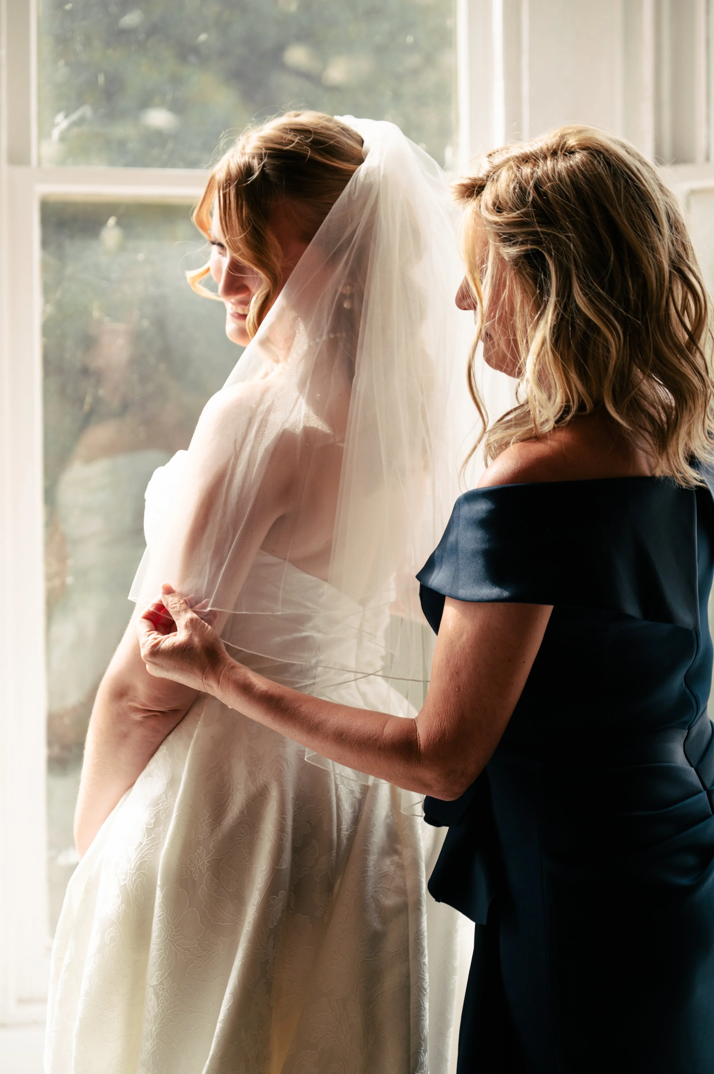 A bride with light brown, wavy hair wearing a white wedding dress and veil, smiling as she looks down; a woman in a dark off-shoulder dress assisting her, standing near a window with natural light.