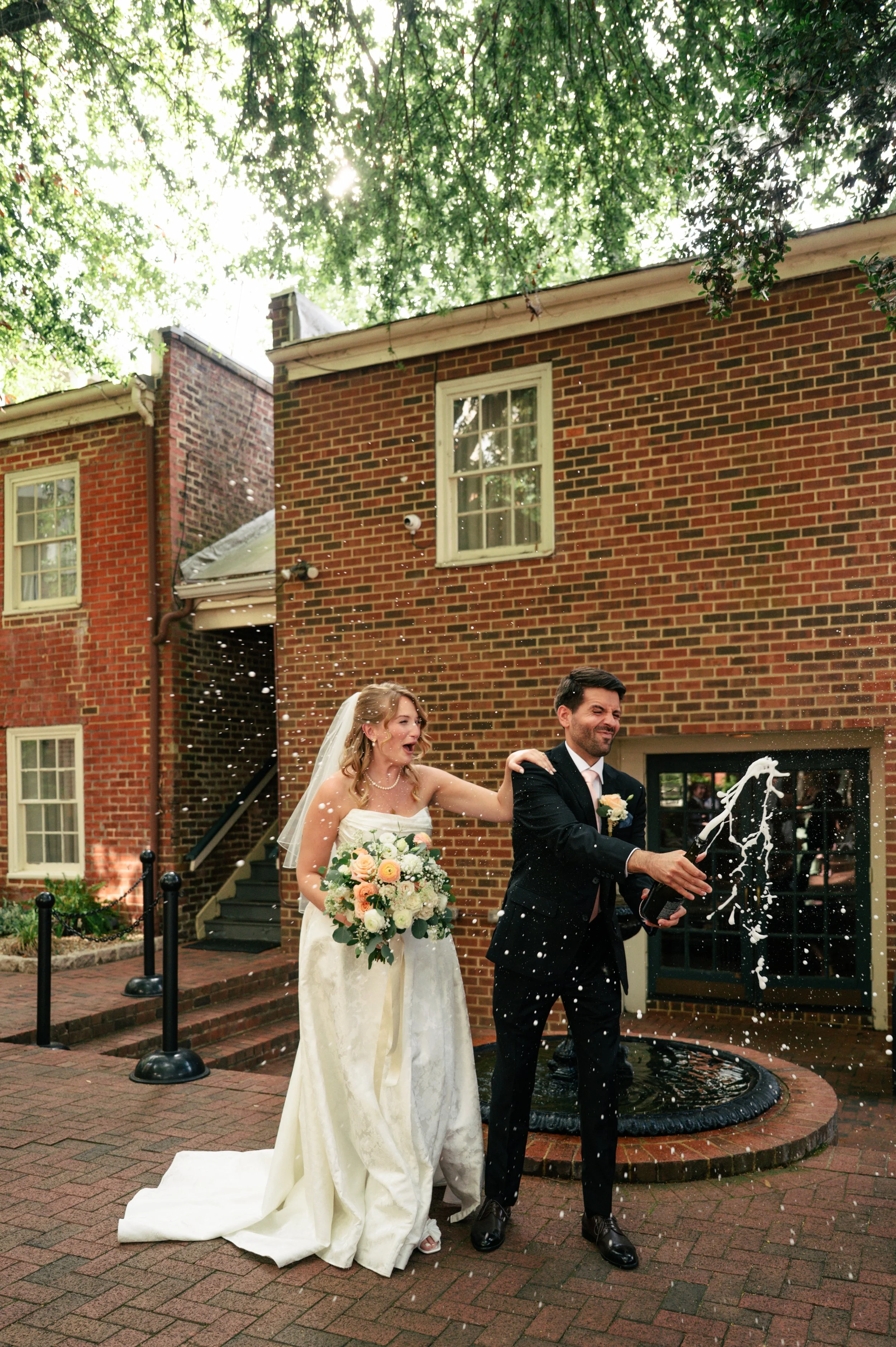 A bride in a white wedding gown holding a bouquet of flowers and a groom in a black suit celebrating outside a brick building after their wedding, with champagne being sprayed.