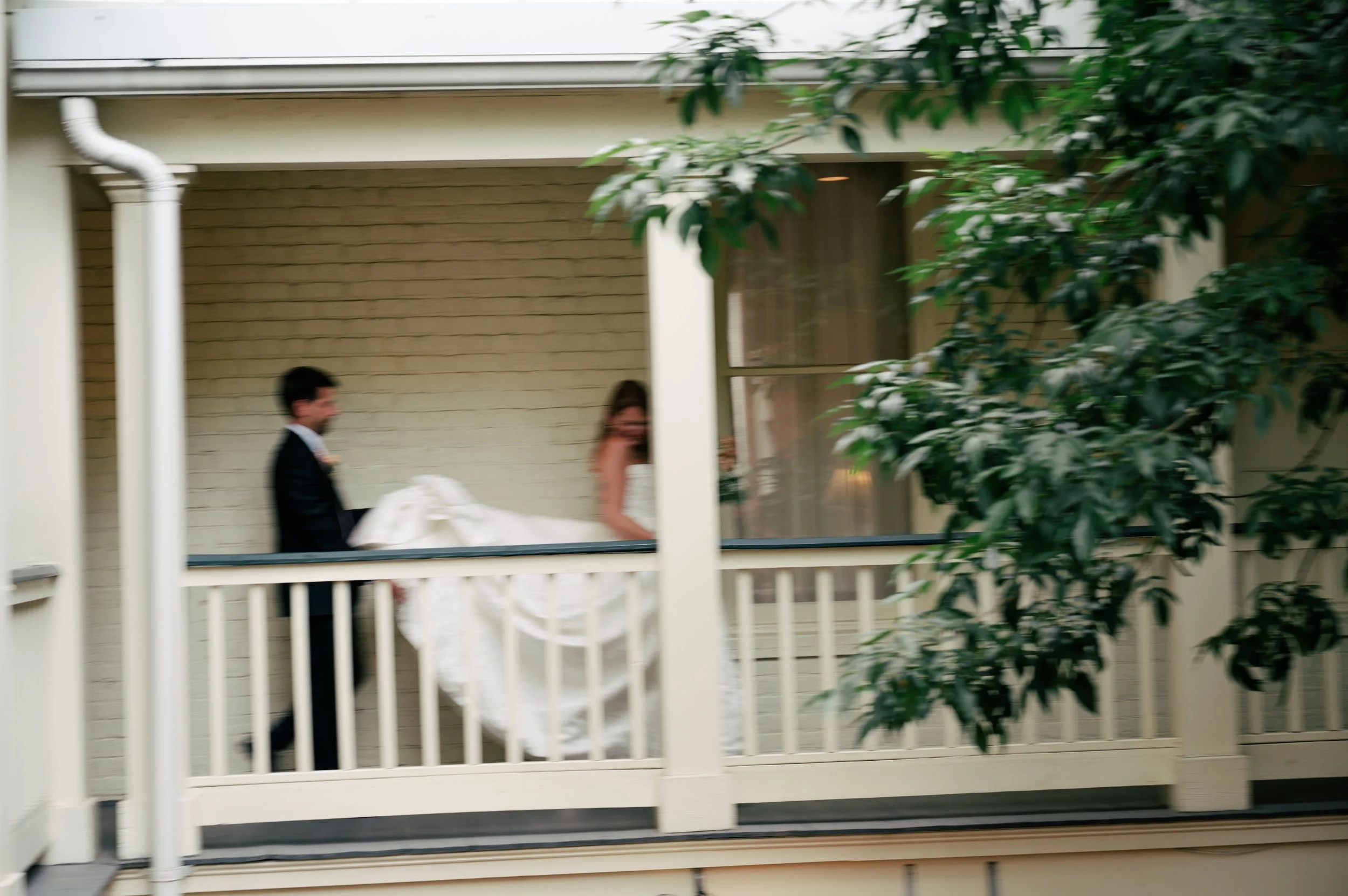 A bride and groom walking on a porch, seen through a window or screen, with green trees partially covering the view.