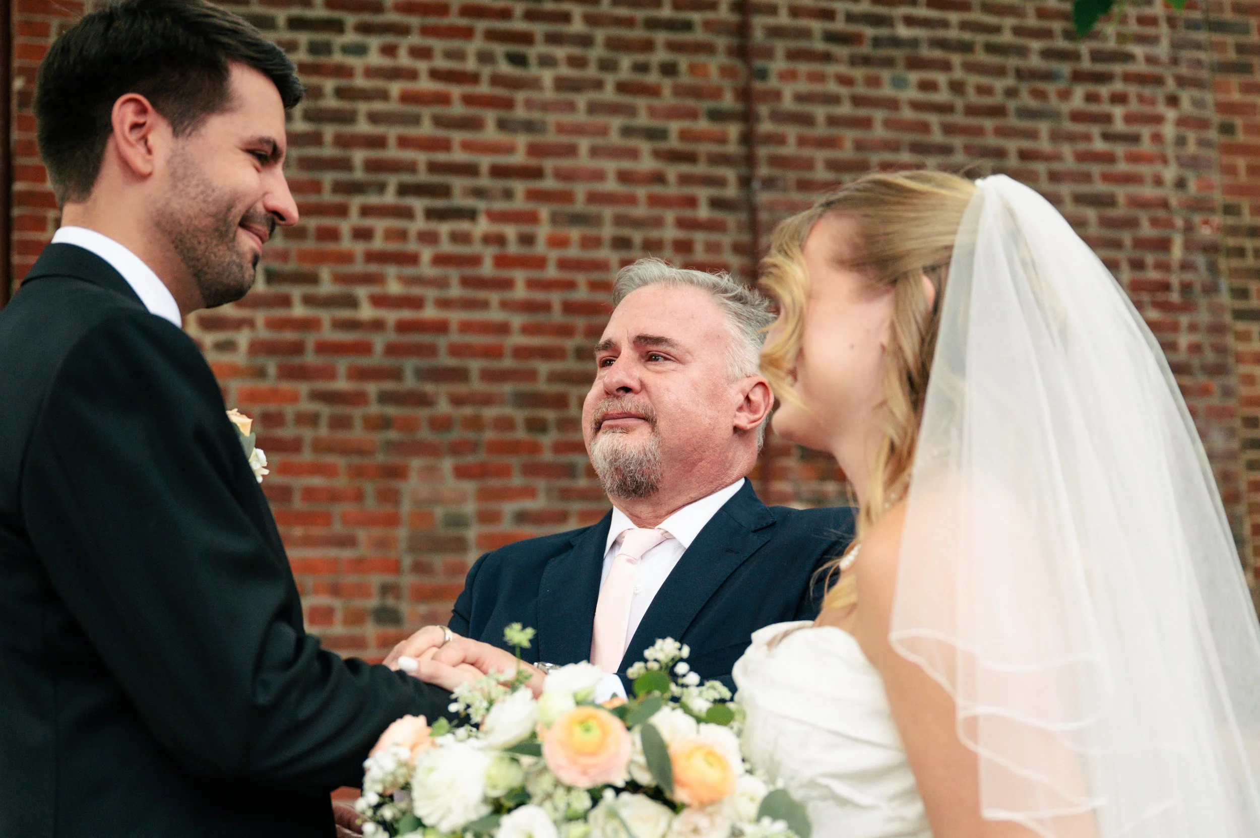 A bride and groom holding hands and facing each other during a wedding ceremony, with a her father looking on, in front of a brick wall.