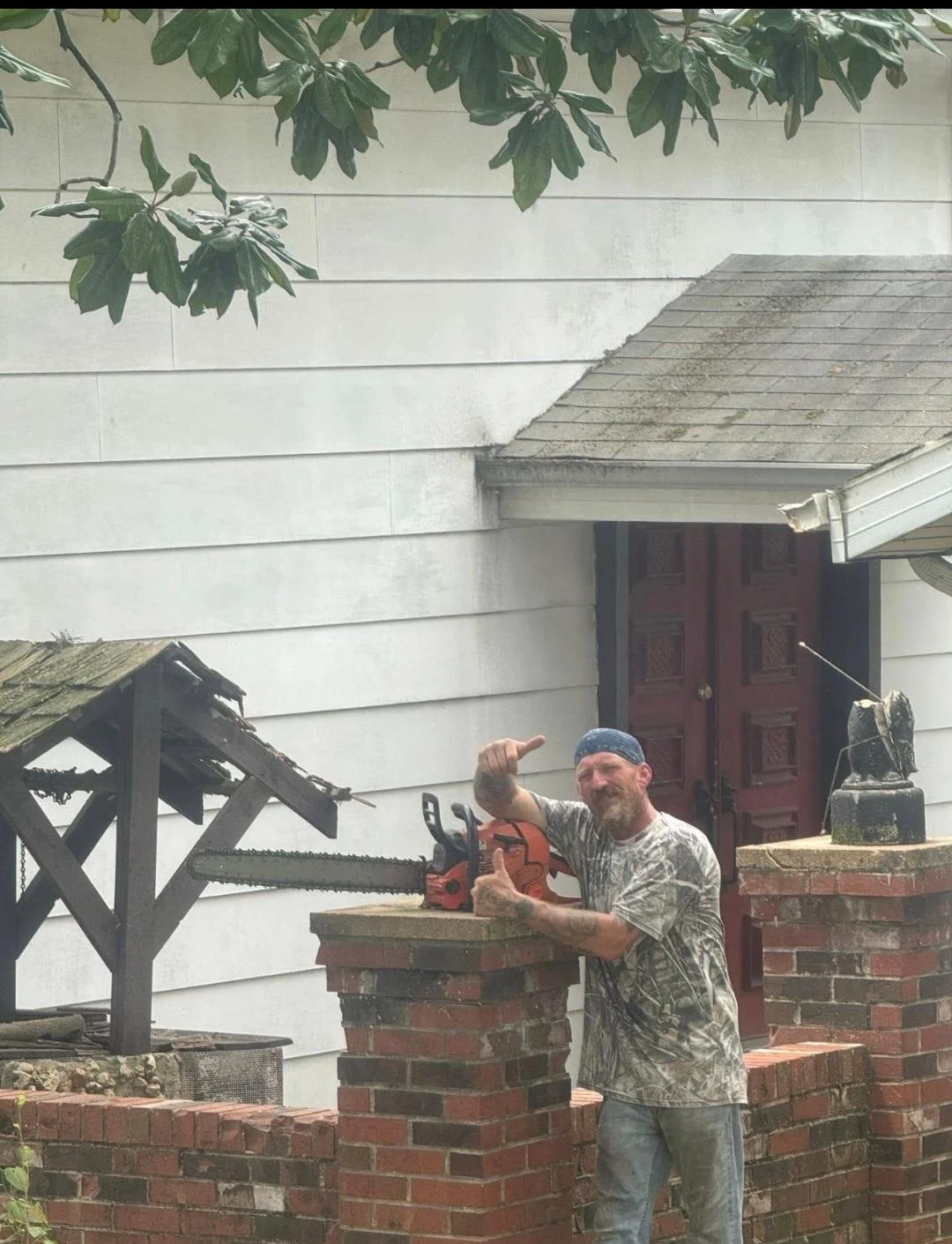A man with a beard and tattoos, wearing a camouflage shirt and blue bandana, stands behind a brick wall with a chainsaw on top, giving a thumbs-up and pointing at it. The background shows a white house wall, a red door, and a decorative eagle sculpture.