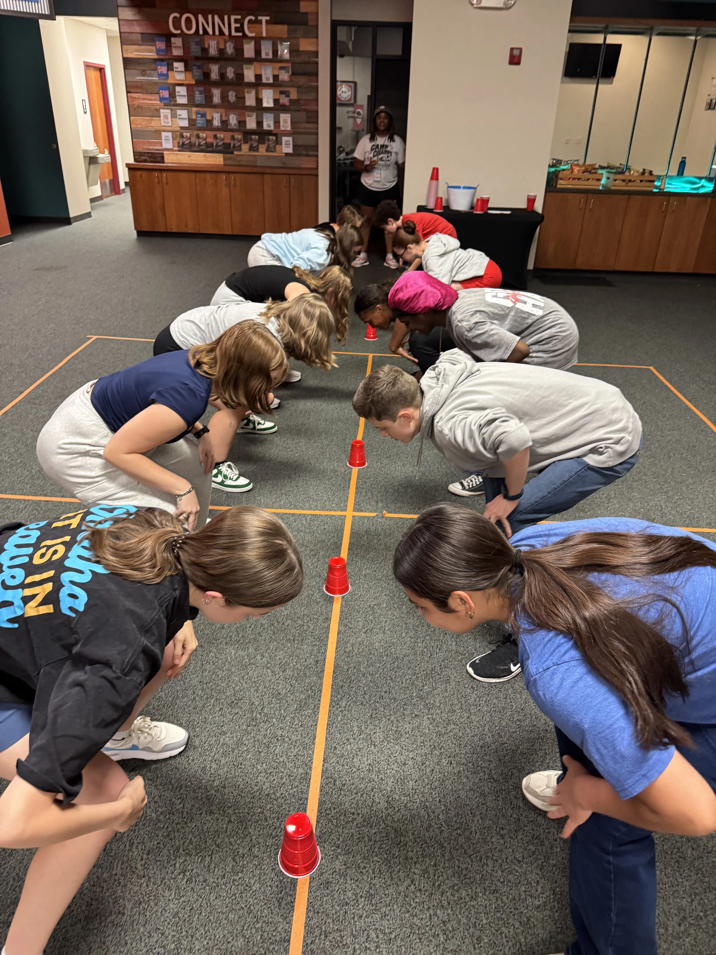 Group of children and teenagers participating in a cup stacking activity on the floor of an indoor space, with orange lines marking the activity area and red plastic cups stacked in the middle.