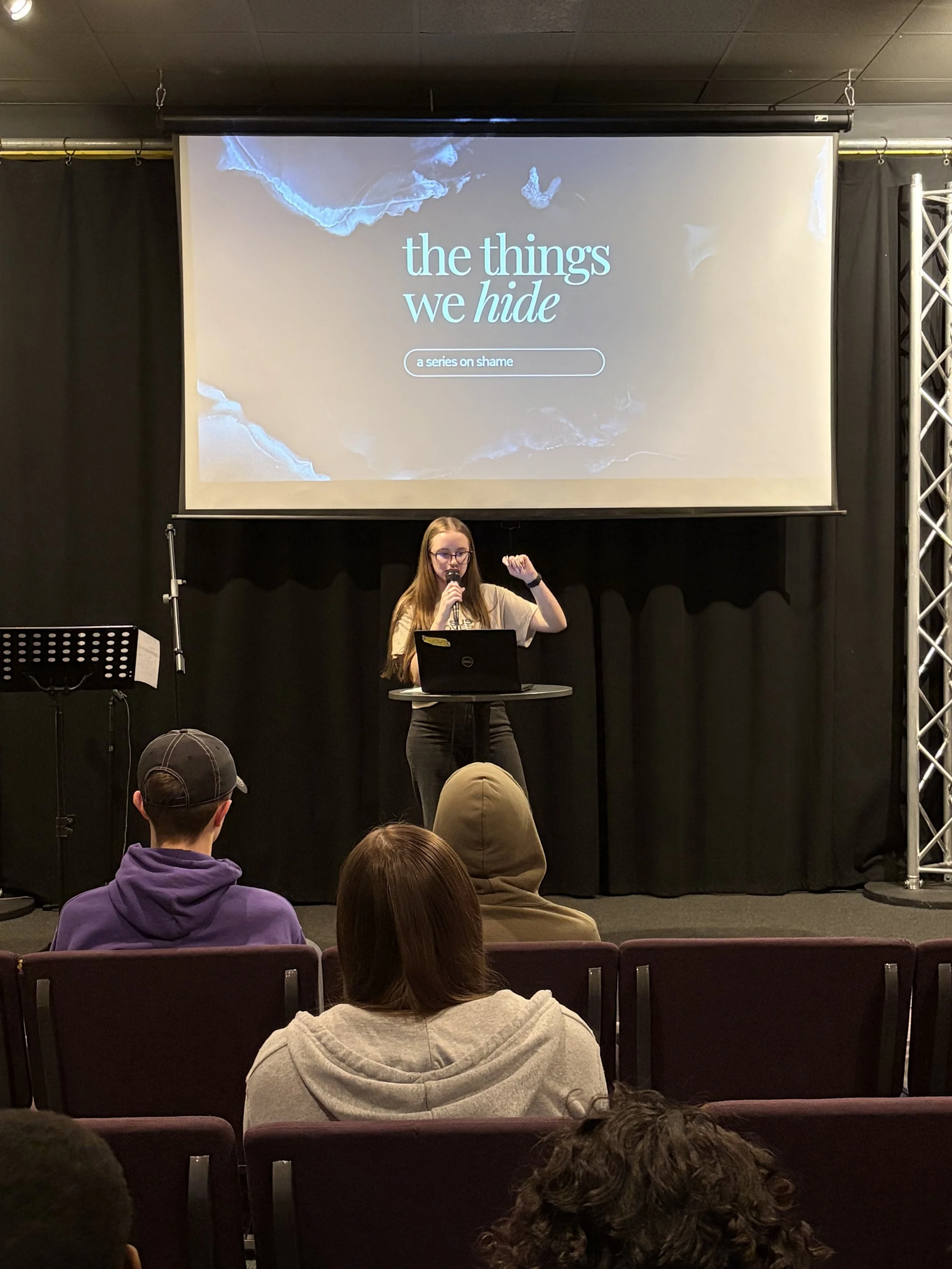 A woman standing at a small round table with a laptop and microphone, giving a presentation in front of a large screen with the title 'the things we hide' and subtitle 'a series on shame'. She is gesturing with her right hand while speaking.