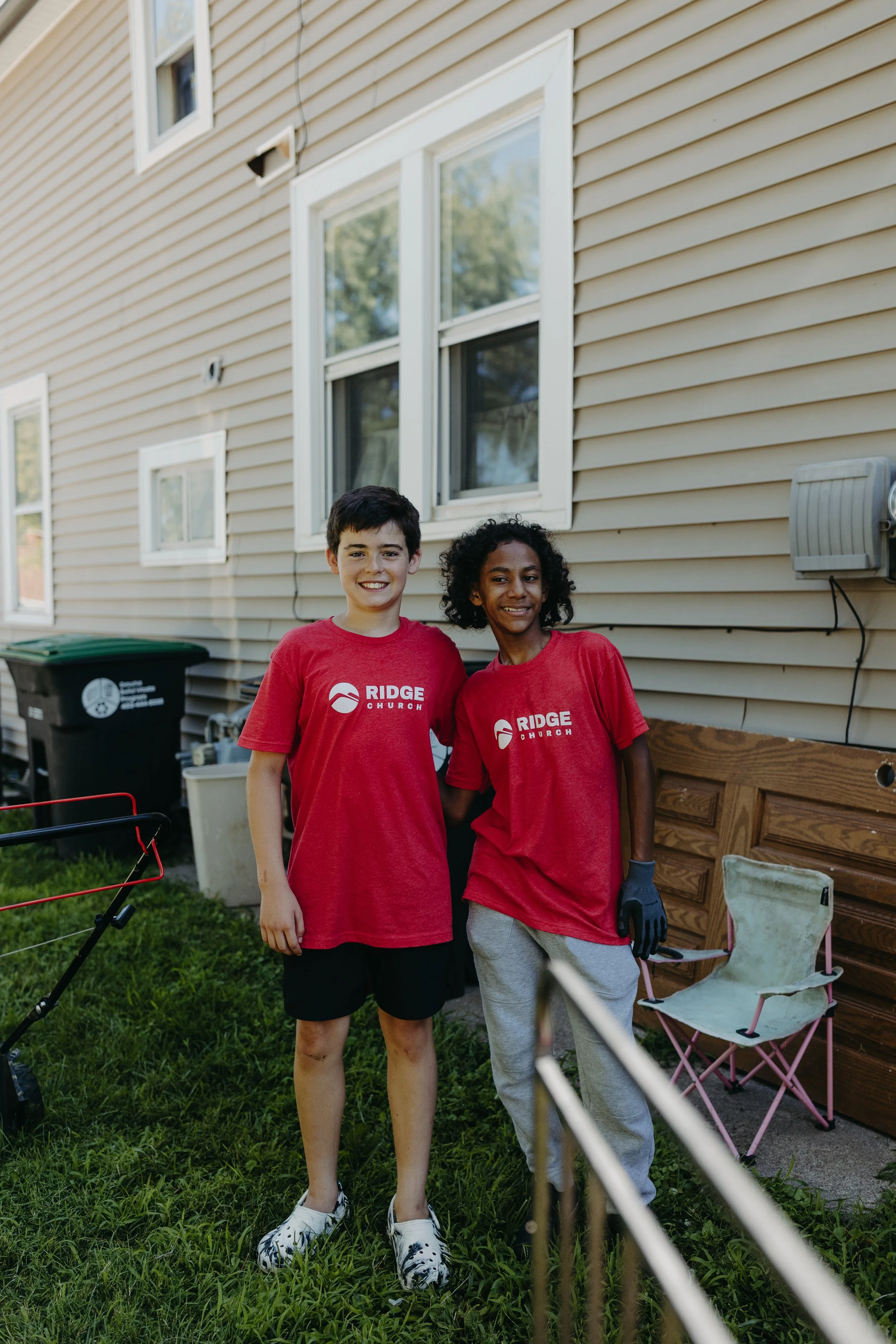 Two children, a boy and a girl, are standing outside a house, wearing red shirts with the words "RIDGE CHURCH". The boy has short dark hair and is smiling, while the girl has curly black hair and is holding a gardening glove. They are standing on gra