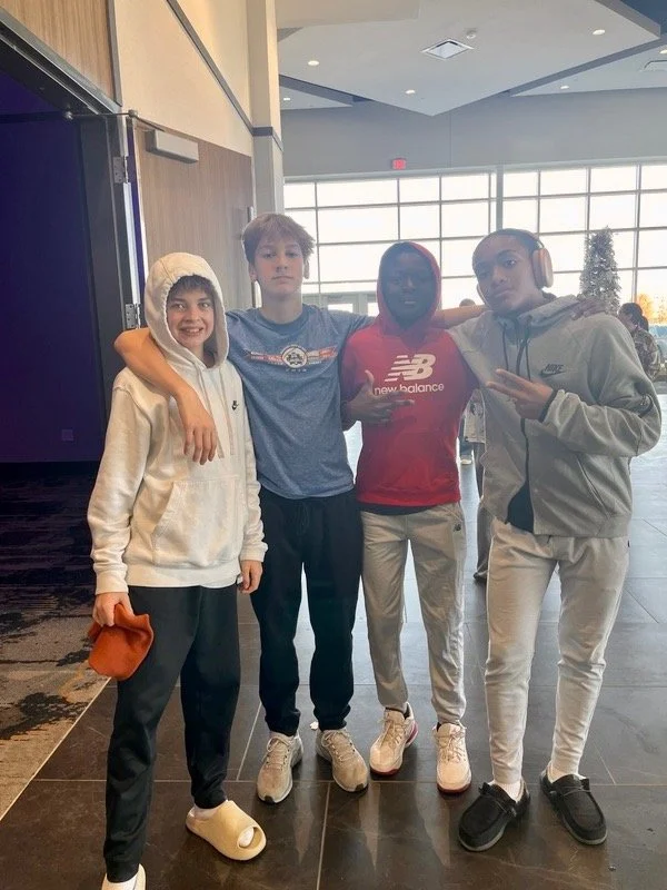 Four young boys standing together indoors with large windows in the background. They are dressed casually in hoodies, sweatpants, and sneakers, and some are wearing headphones and a hoodie with the hood up. They appear to be friends posing for a phot