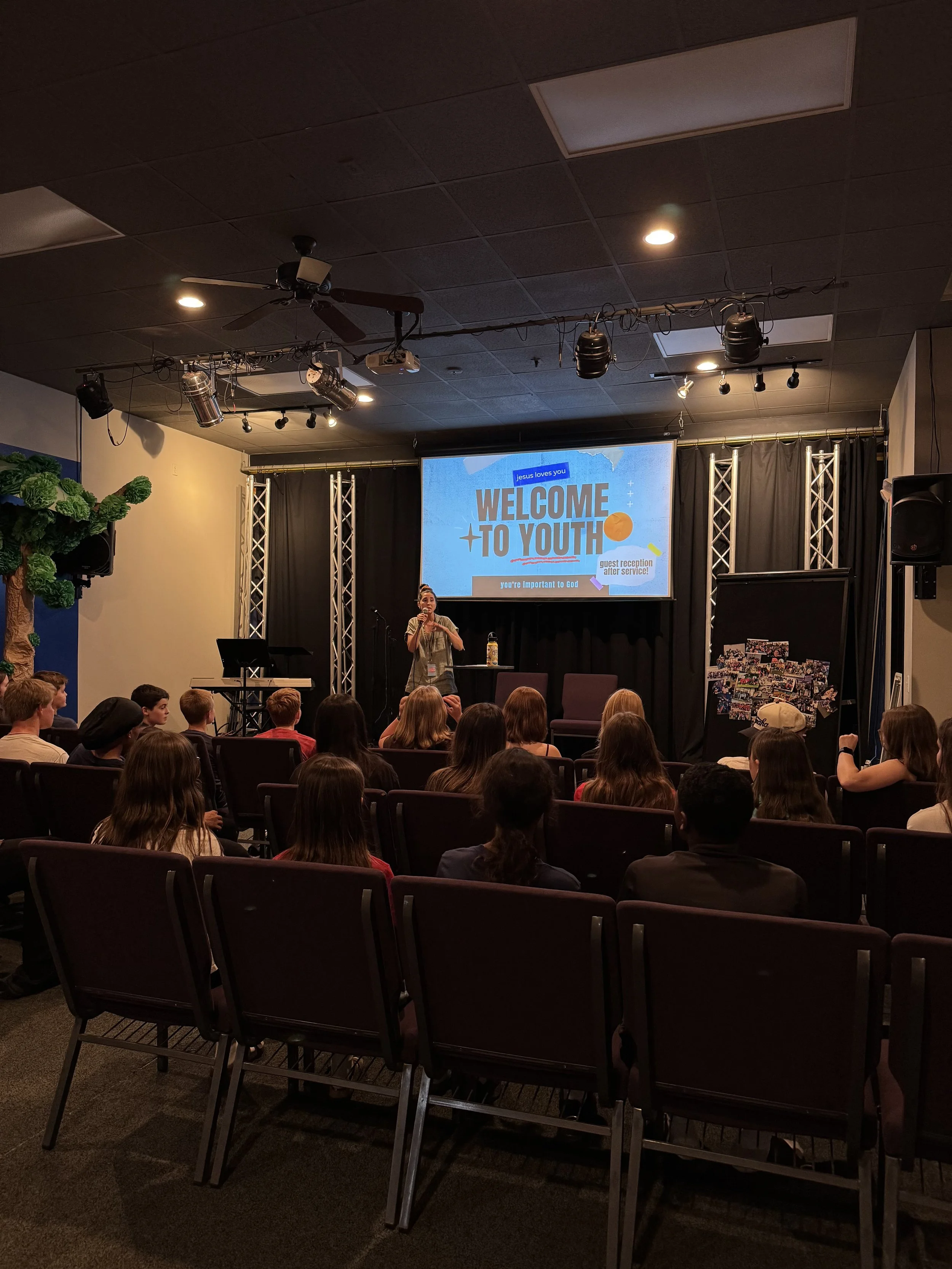 A church youth group event with a stage, a large screen displaying a welcome message, a woman speaking at a microphone, and several young attendees seated in chairs.