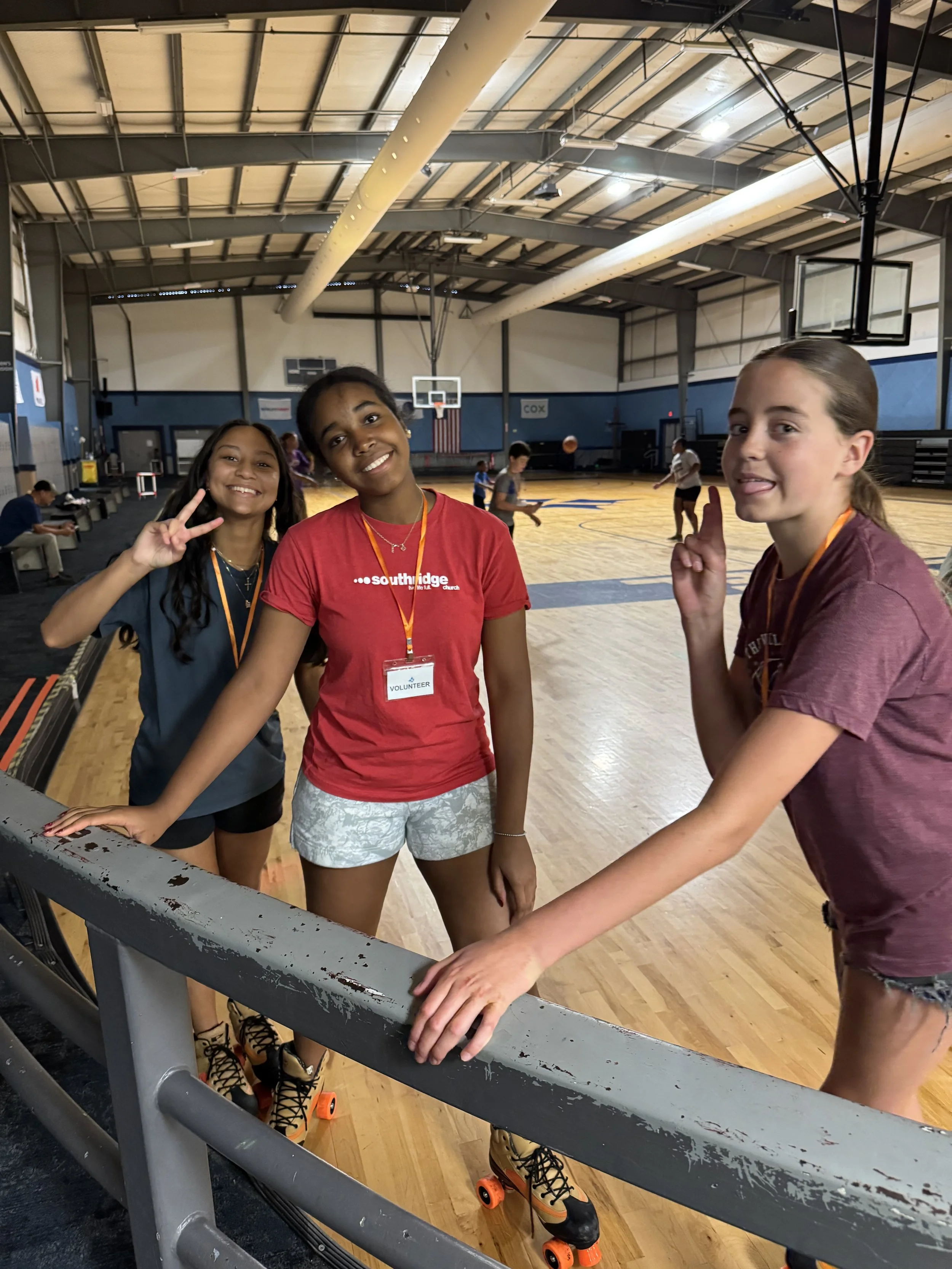 Three smiling young girls with roller skates posing in a gymnasium with a basketball court, two of them showing friendship signs and one sticking out her tongue, while a boy and other children are in the background.
