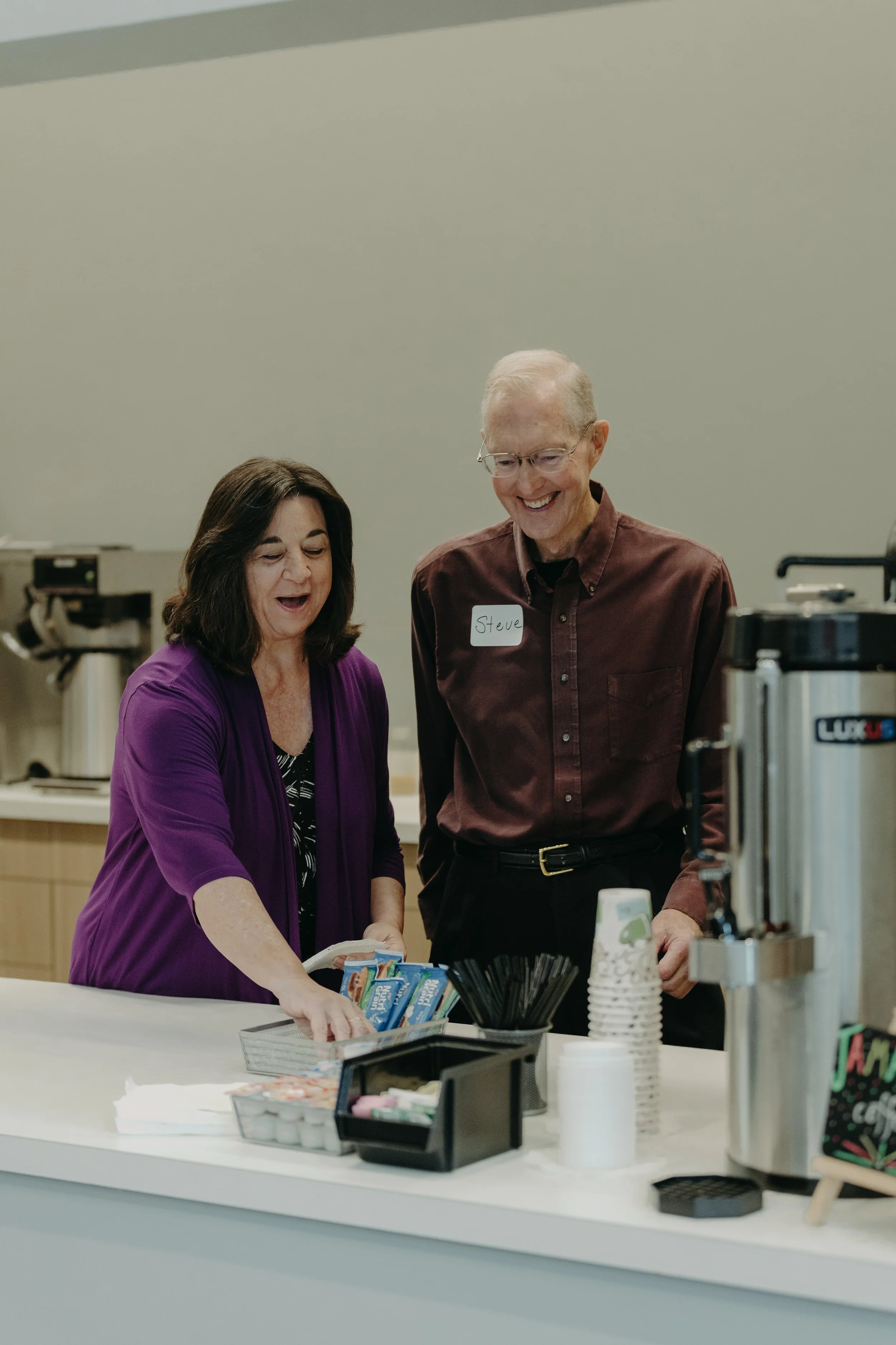 A woman in a purple jacket and a man with a name tag that says "Steve" are smiling and looking at items on a counter, possibly in a break room or kitchen area.