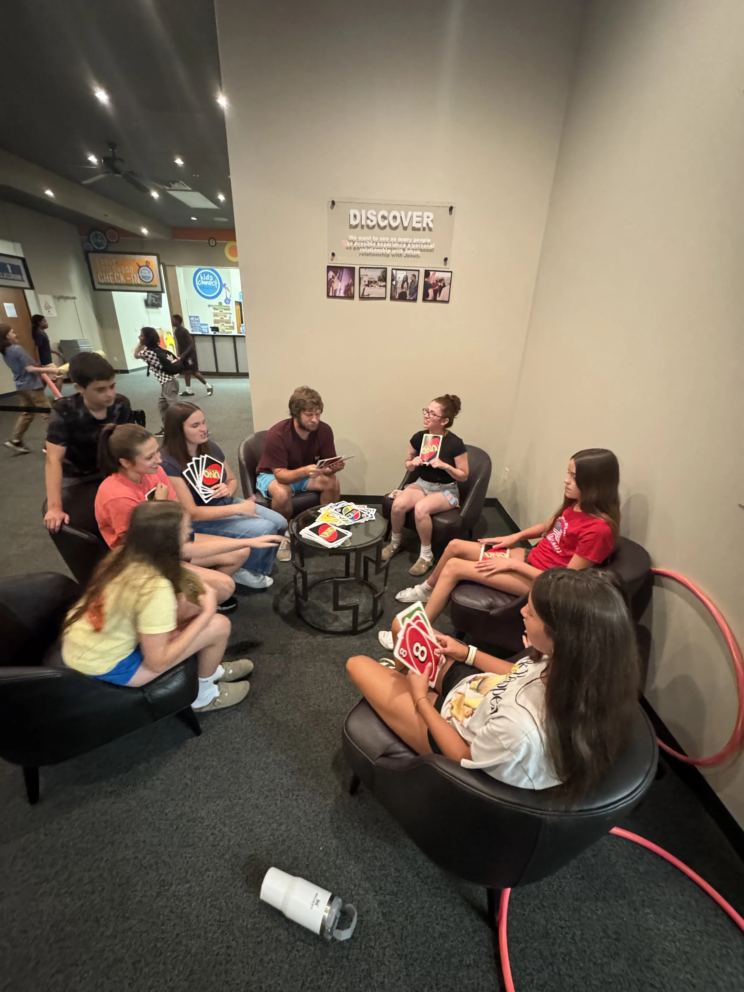 A group of young people sitting on black sofas in a circle, holding UNO cards, in an indoor setting with other people walking in the background.