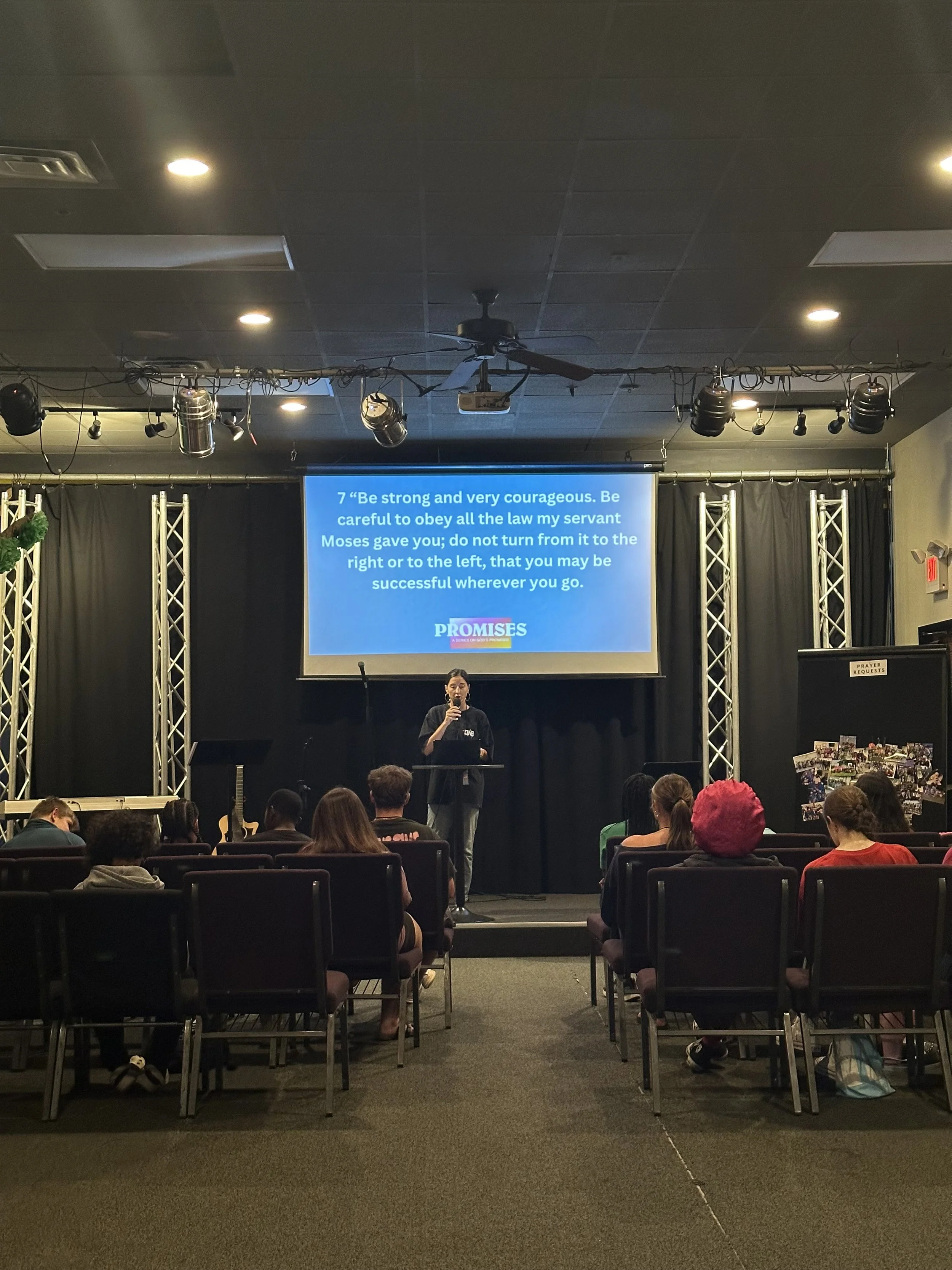 People sitting in rows of chairs facing a stage with a woman speaking at a podium. Behind her is a large screen displaying a Bible verse from Numbers 21:17 about being strong, courageous, and obedient. The stage is decorated with metal trusses, curta