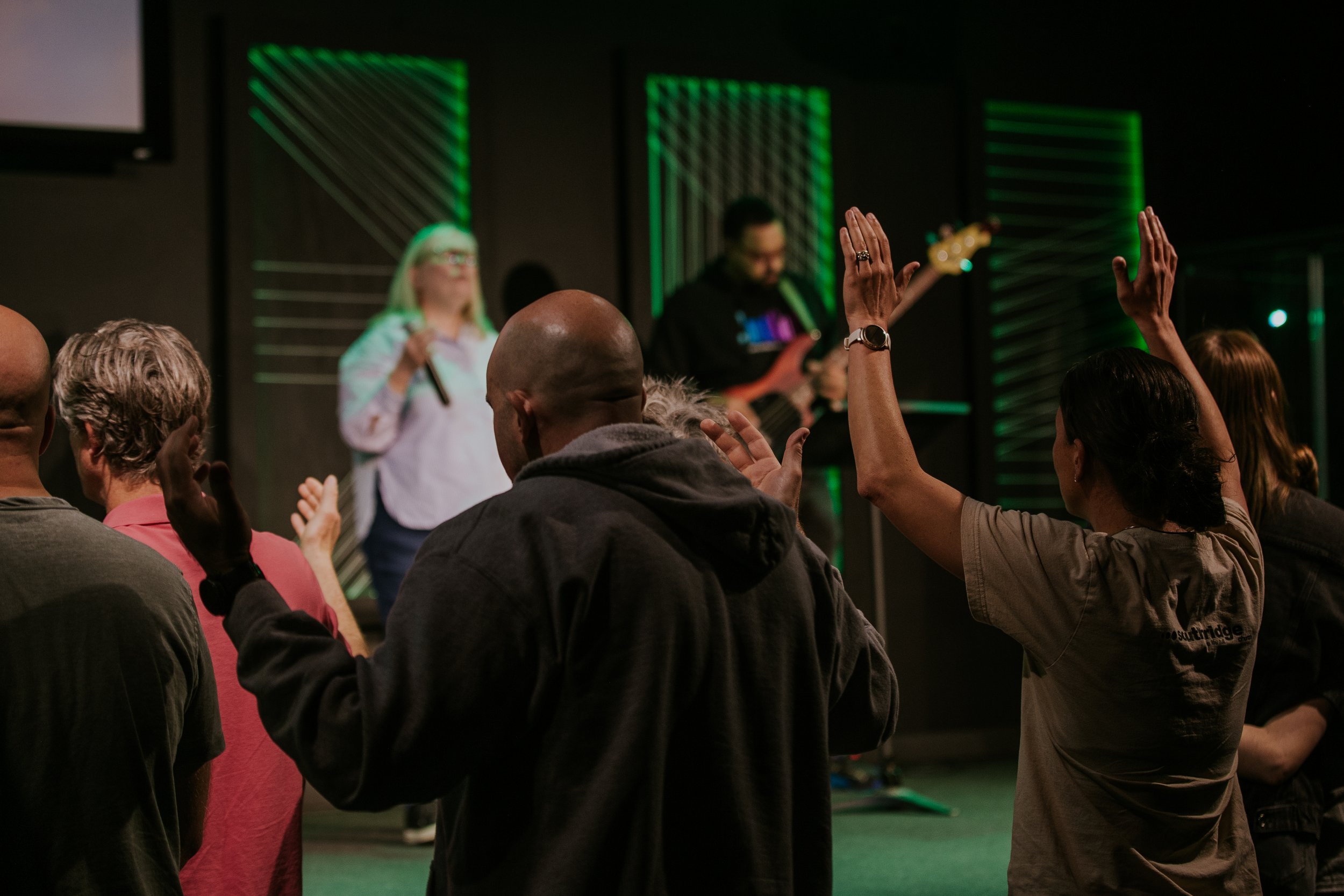 A diverse group of people participating in a live event or gathering, with some individuals raising their hands and others facing a stage where a woman is speaking.