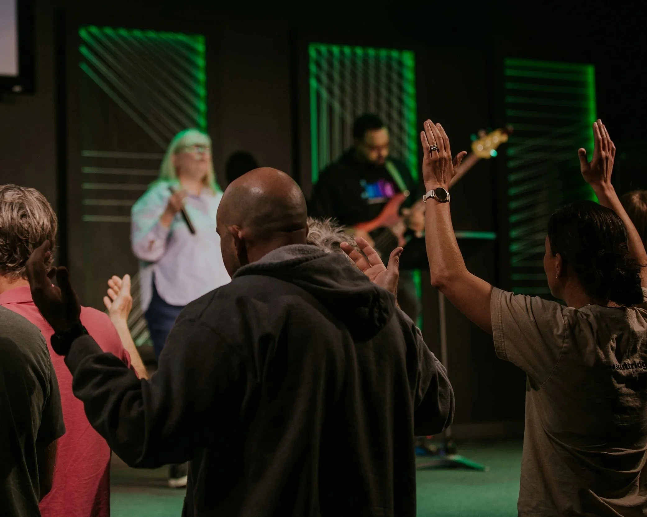 People with raised hands during a presentation or event in a dark room with green backlit panels.