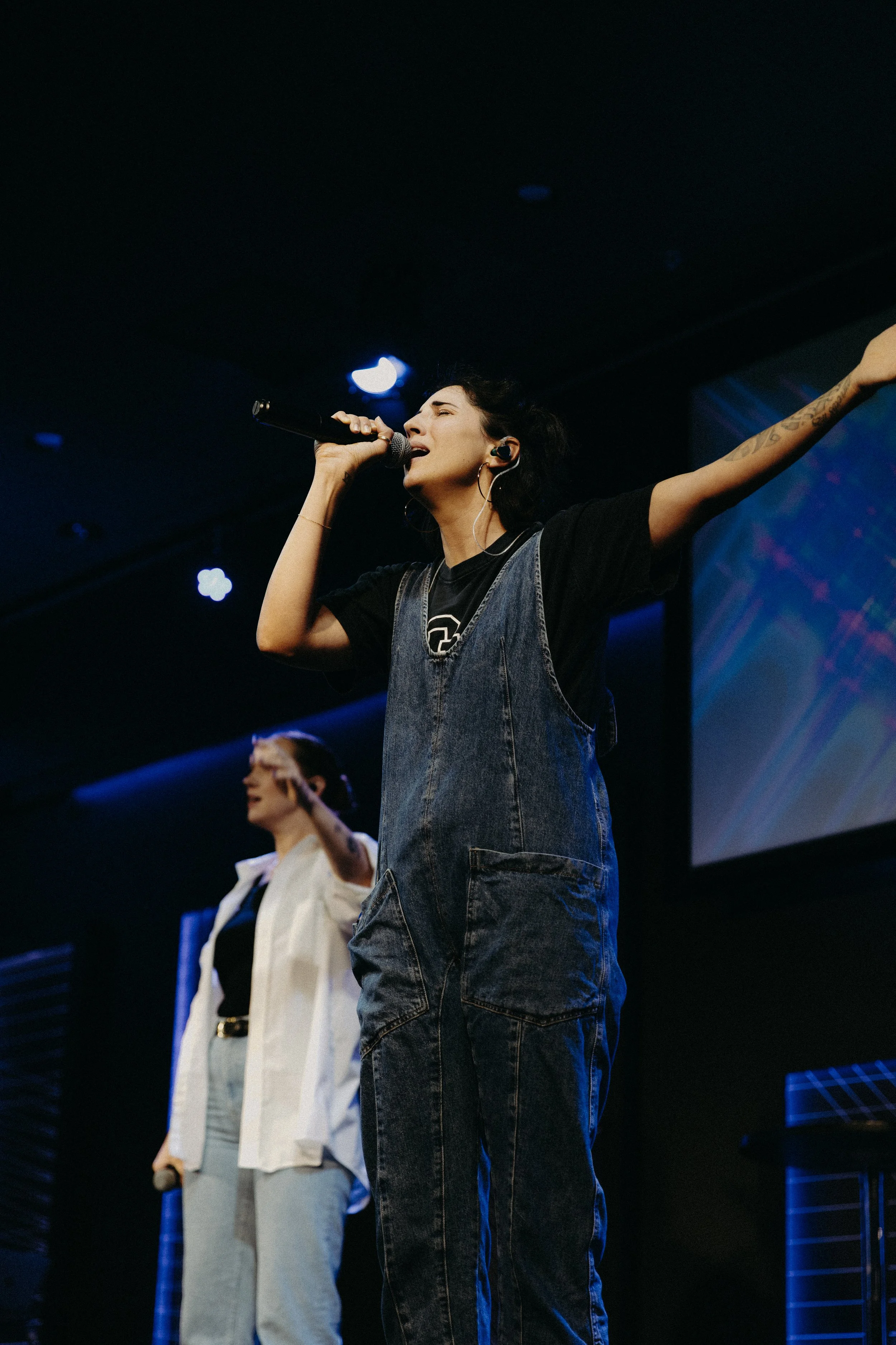 A woman singing into a microphone on stage with her arm raised, wearing a black t-shirt and denim overalls, with another woman in the background.