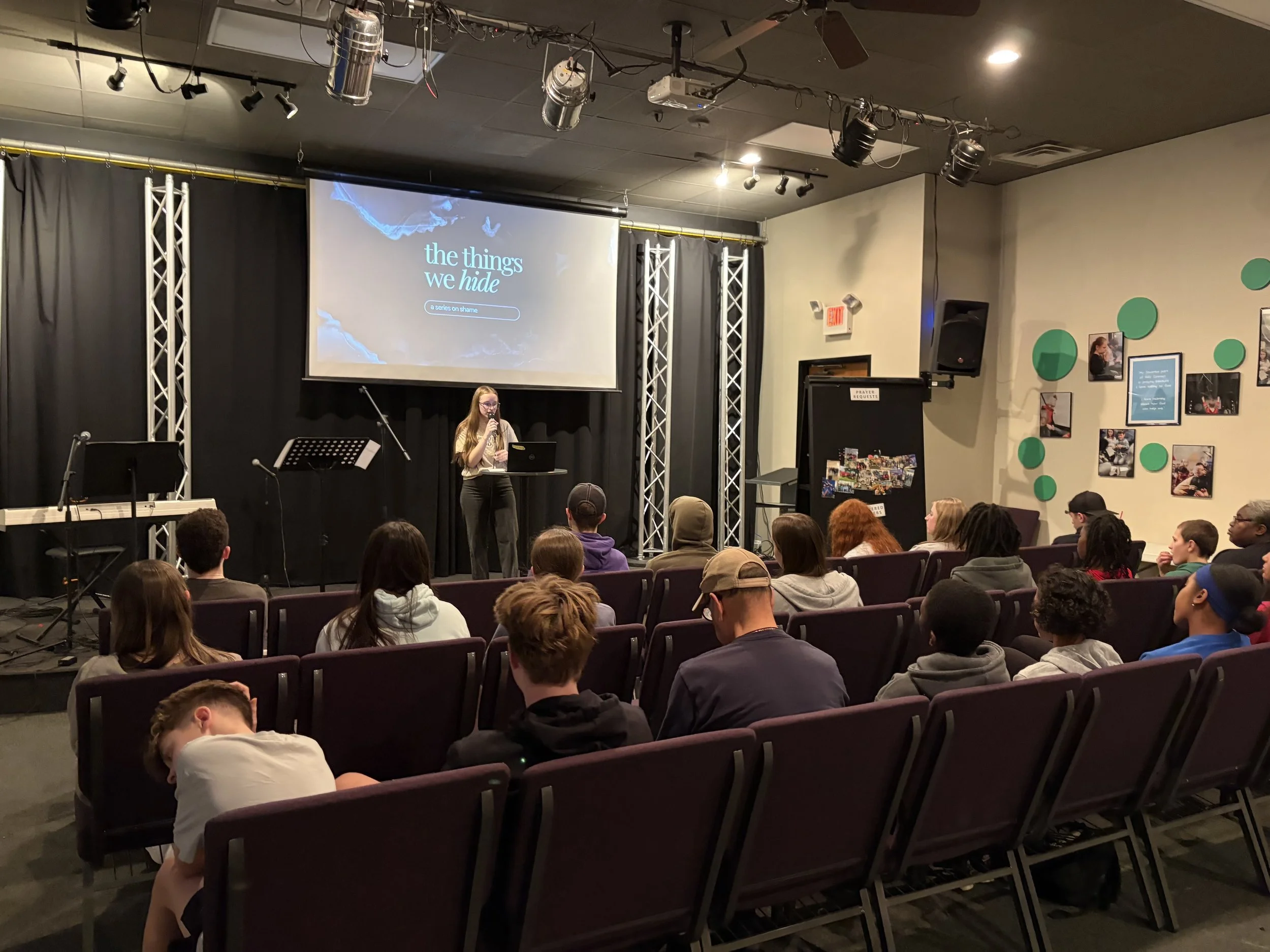 A church or community center auditorium with a stage where a young woman is speaking into a microphone. A large screen behind her displays the title 'the things we hide' and the subtitle 'a series on shame.' Audience members are seated, listening att