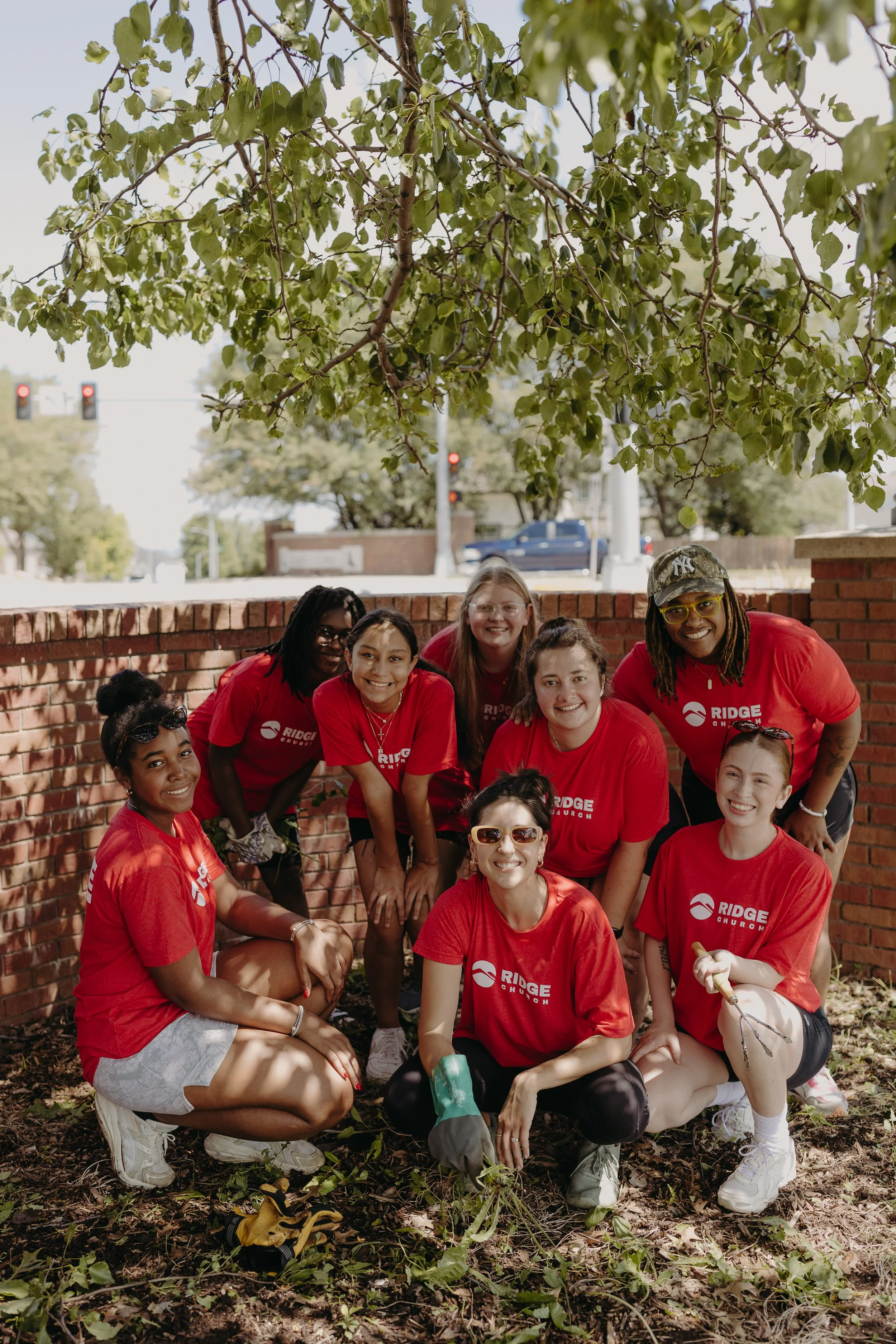 A group of diverse young people wearing red Ridge Church T-shirts gathered outdoors, smiling and posing for a photo during a community service project or event under a tree.