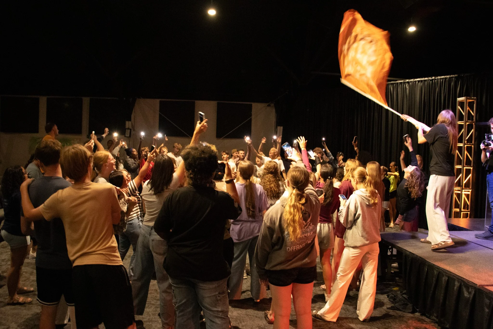 A woman on stage waving an orange flag to a crowd at a concert or event in a dark indoor venue. The audience is taking photos and videos with their phones.