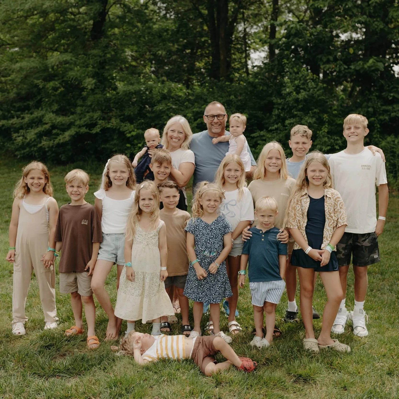 A large group of children and two adults standing outdoors on grass with trees in the background, posing for a photo.