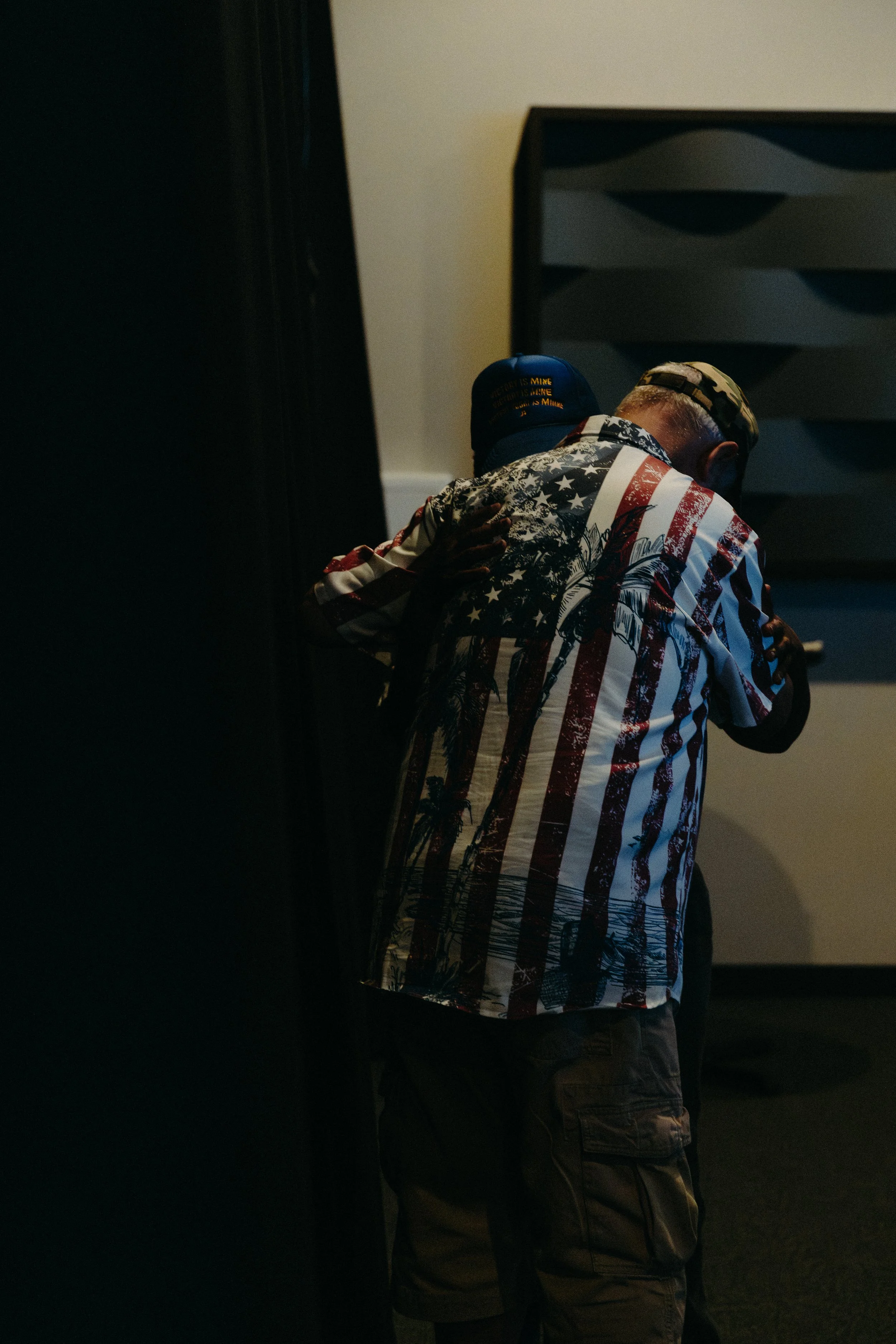 A man with a camouflage cap and a patriotic American flag-themed shirt, seen from behind, standing and leaning forward in a dimly lit room, with part of a black cabinet on the left and a light-colored dresser with drawers on the right.