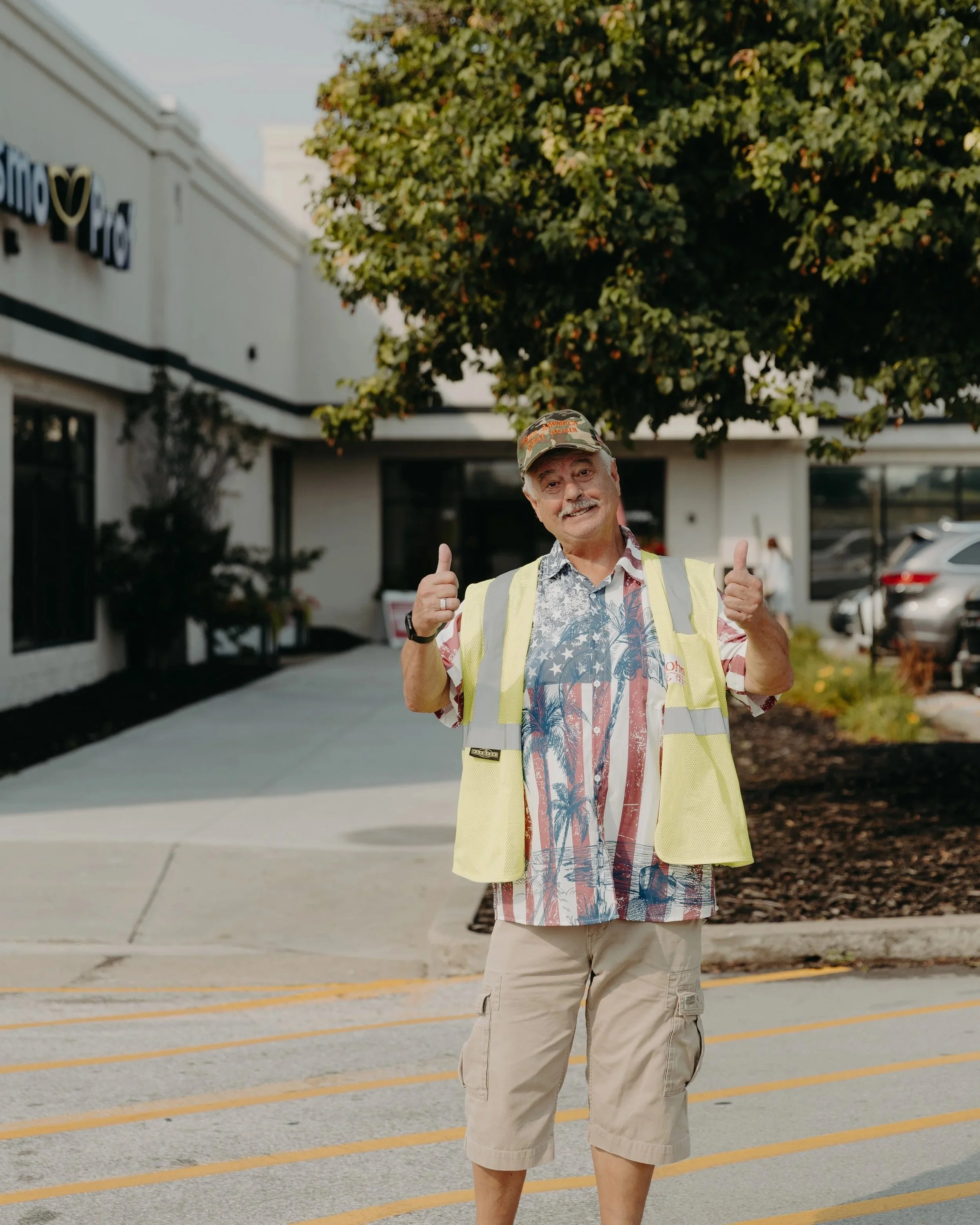 A man smiling and giving two thumbs up outside a shopping center, wearing casual clothes including a Hawaiian shirt, beige shorts, a yellow safety vest, and a cap. There are cars parked in the background, a large green tree, and a building with a partially visible sign.