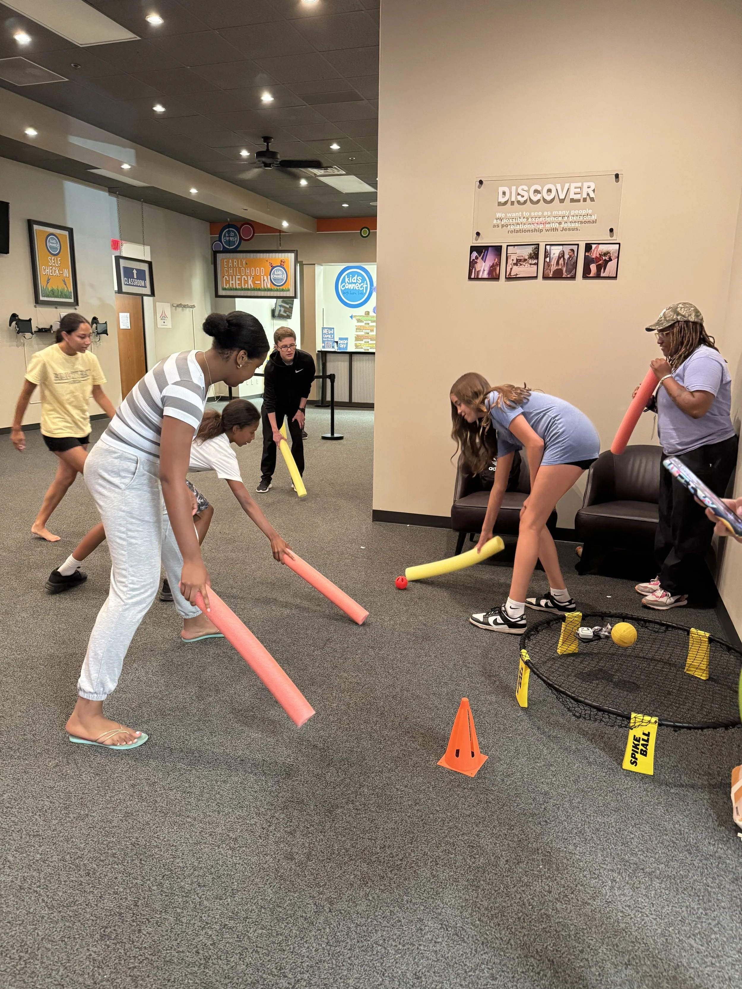 Young people playing hockey in an indoor space. They are using foam sticks and a ball, with some wearing casual clothing and others in athletic attire. Cones and a small goal with balls are on the floor, and signs on the wall indicate early childhood