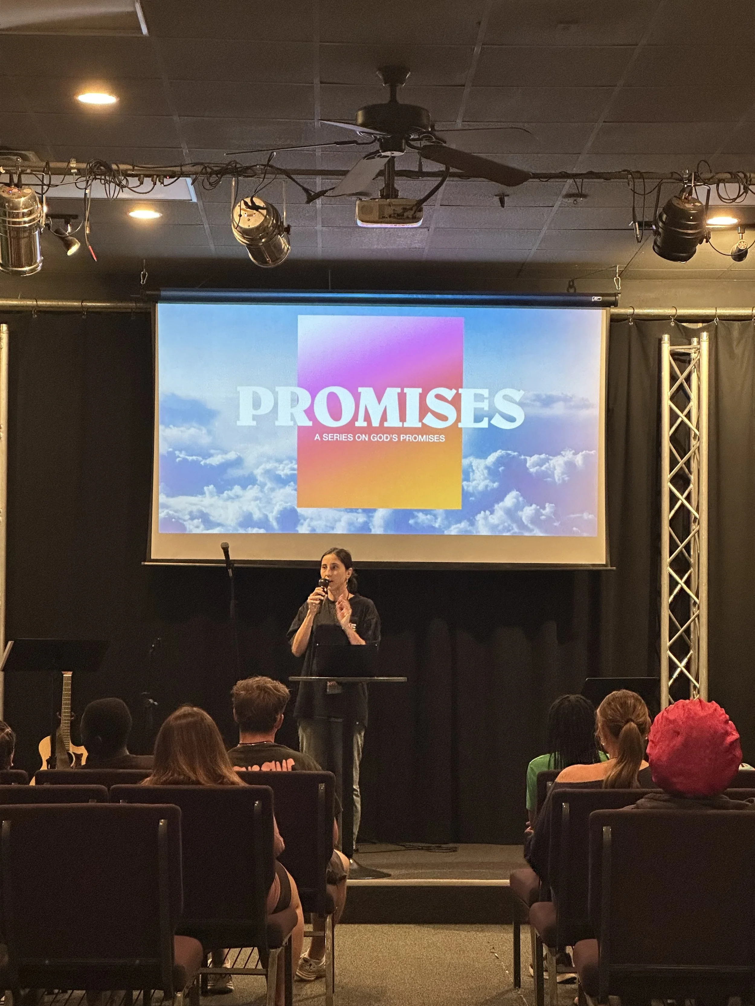 A woman speaking at a podium during a presentation titled 'Promises: A Series on God's Promises,' with an audience seated in front of her. A large screen behind her displays the title and a background of sky and clouds.